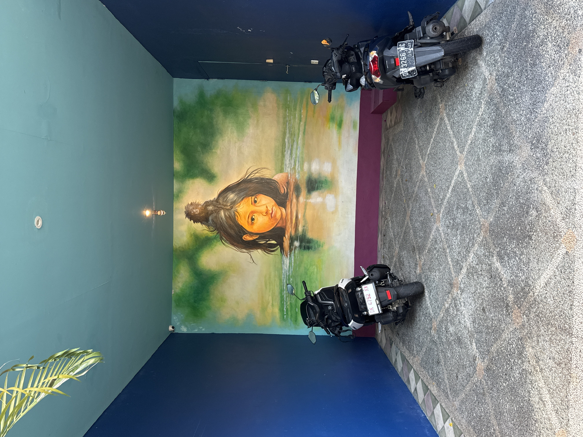 The image shows a small outdoor parking area in Kuta, Indonesia. The area is enclosed by walls painted in varying shades of blue and green, with a mural depicting a woman emerging from a body of water on the central wall. Two parked motorbikes are visible on the right, occupying most of the space. The floor is covered in gray paving stones. The lighting appears to be a combination of natural daylight and a small overhead light fixture. A plant is visible on the left side.