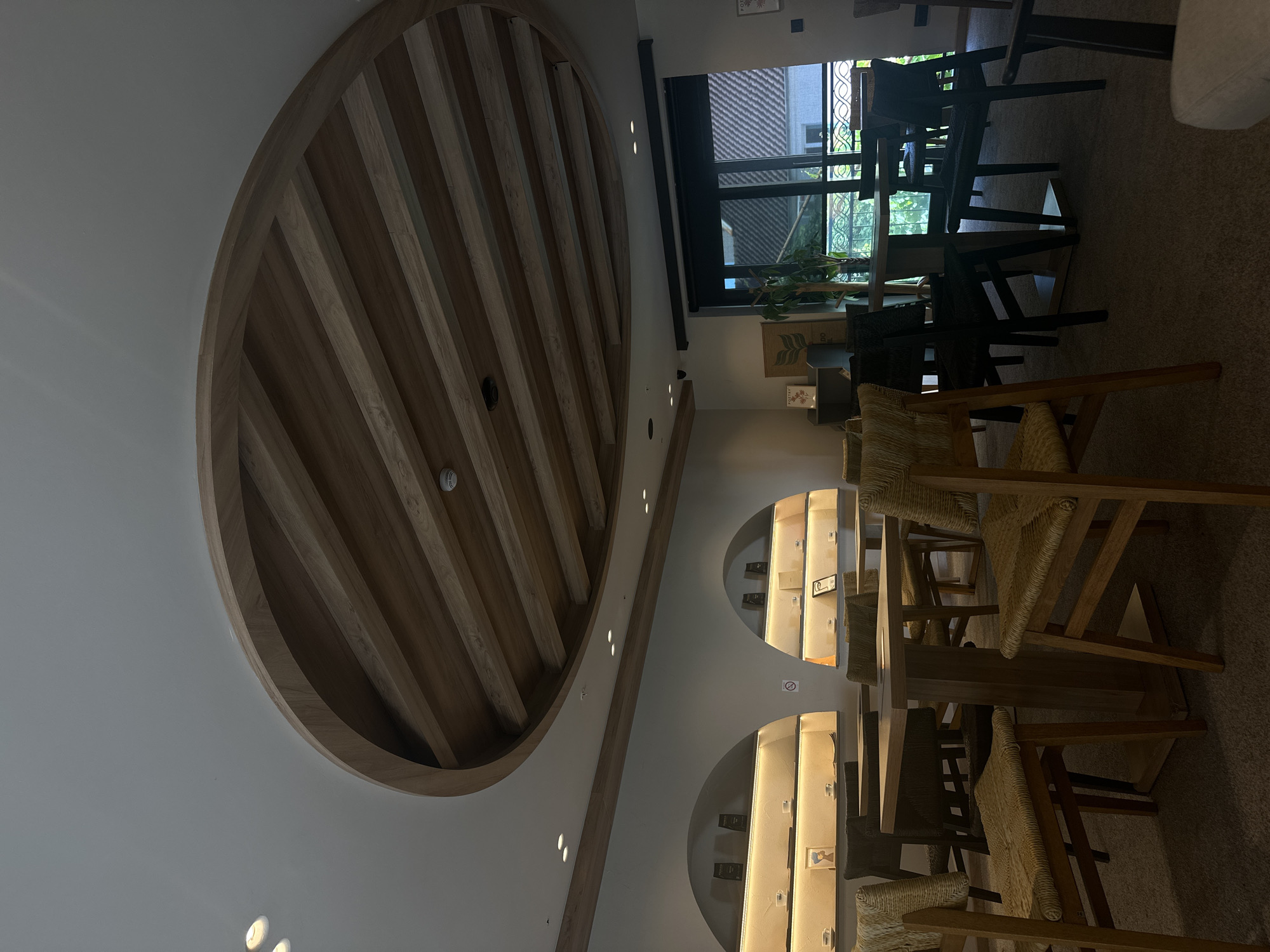 The image shows the interior of a cafe, possibly in Podgorica, Montenegro, though this location is not explicitly verifiable from the image alone.
The scene is a brightly lit interior featuring a prominent, decorative wooden circular ceiling element made of wooden slats. The ceiling is white. Below, the space is furnished with wooden tables and chairs, a combination of dark wood and light straw-colored seating. The chairs are arranged around the tables, suggesting a dining or seating area. On the right side of the image, a partially visible window reveals some greenery outside.
Additionally, there are arched, illuminated recessed display cases on the wall, likely showcasing merchandise or decorative items. The overall impression is a modern, stylish cafe interior. There are no people visible in the image.