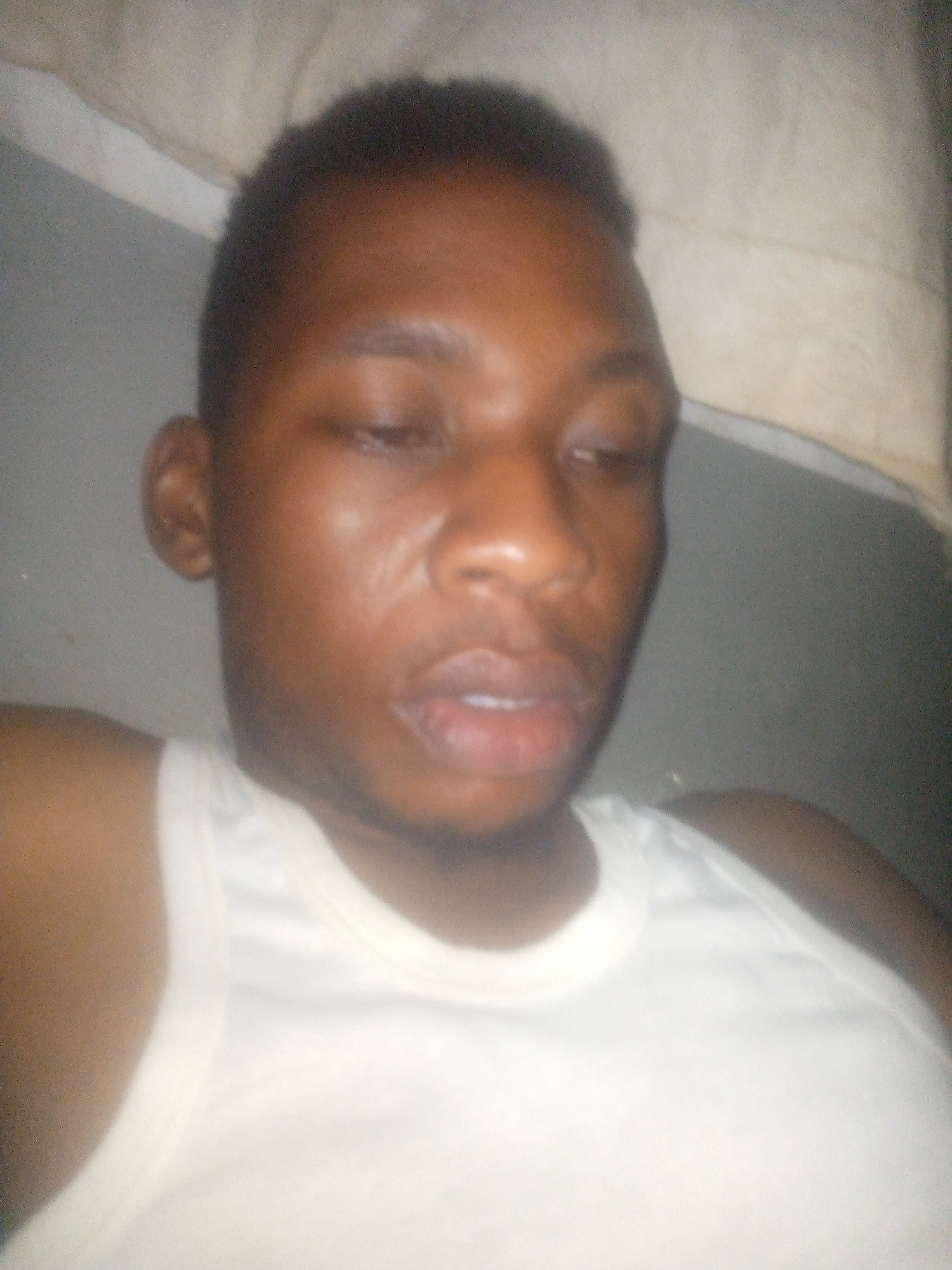 A medium close-up shot shows an African man with closed eyes. He is wearing a white tank top. The background is a gray wall and a white cloth. The location and city are unknown.