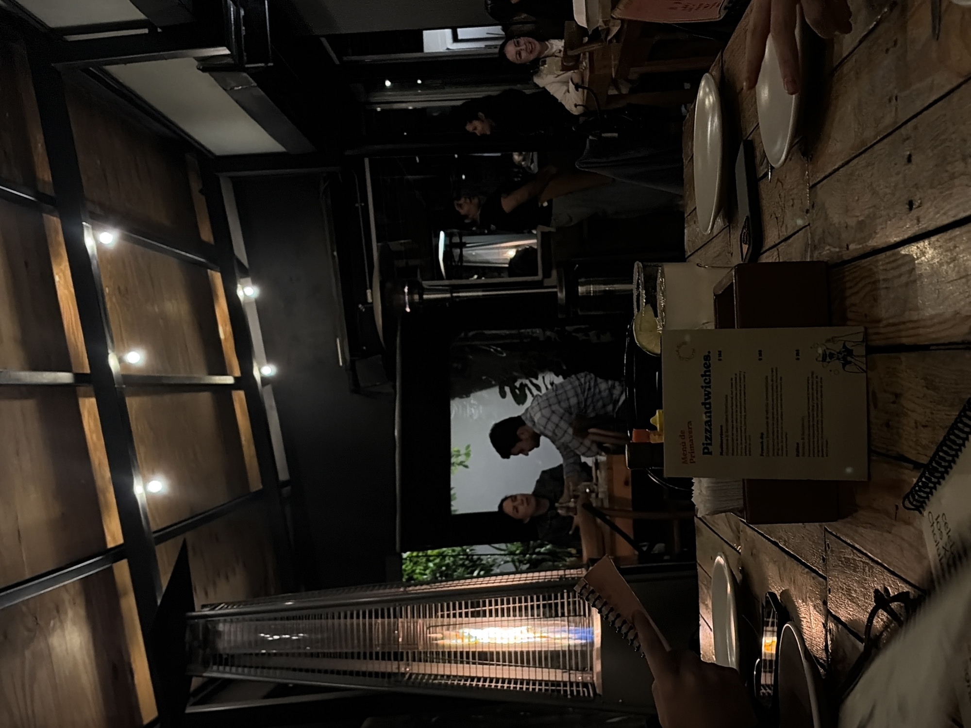 The image shows the interior of a restaurant, seemingly outdoors or semi-outdoors, based on the presence of a patio heater and visible vegetation. There are several people in the background, some seated at tables. In the foreground, a wooden table is set with plates, cutlery, and a menu, indicating that it is ready for diners. A person's hand is visible, possibly holding a notepad or menu. The lighting appears dim and ambient, suggesting an evening setting. The overall scene conveys a relaxed, dining atmosphere, although the specific location within the city of Unknown, Unknown, is not directly discernible from the image.