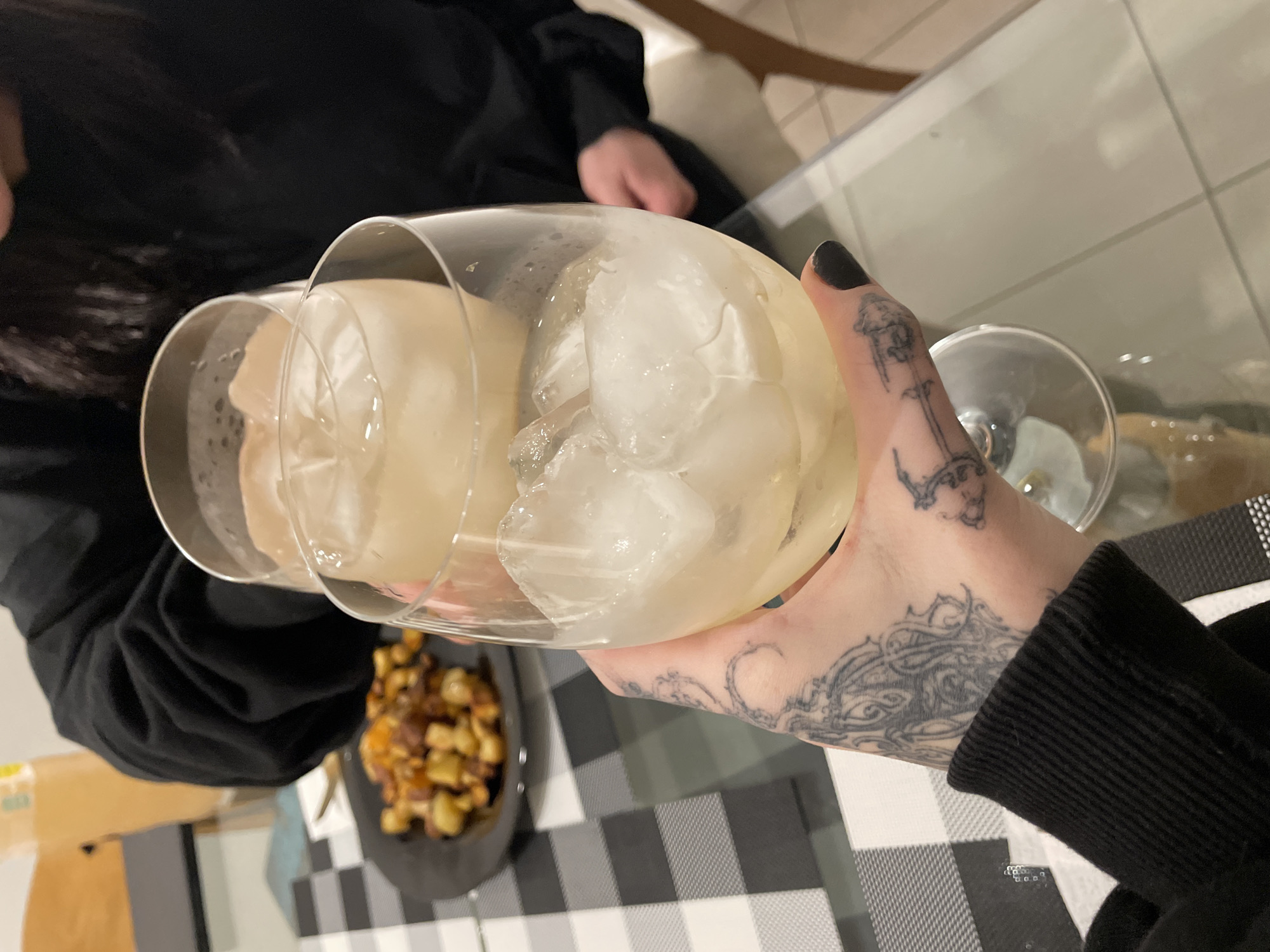 A person is holding two glasses filled with ice and a clear liquid, presumably a beverage. The person's hand has tattoos and is wearing a black sweater. A plate of food, possibly potatoes, is visible on the table. The table has a black and white checkered tablecloth. The environment suggests a casual dining setting indoors. The location in the city of Buenos Aires, Argentina, cannot be determined from the given image.