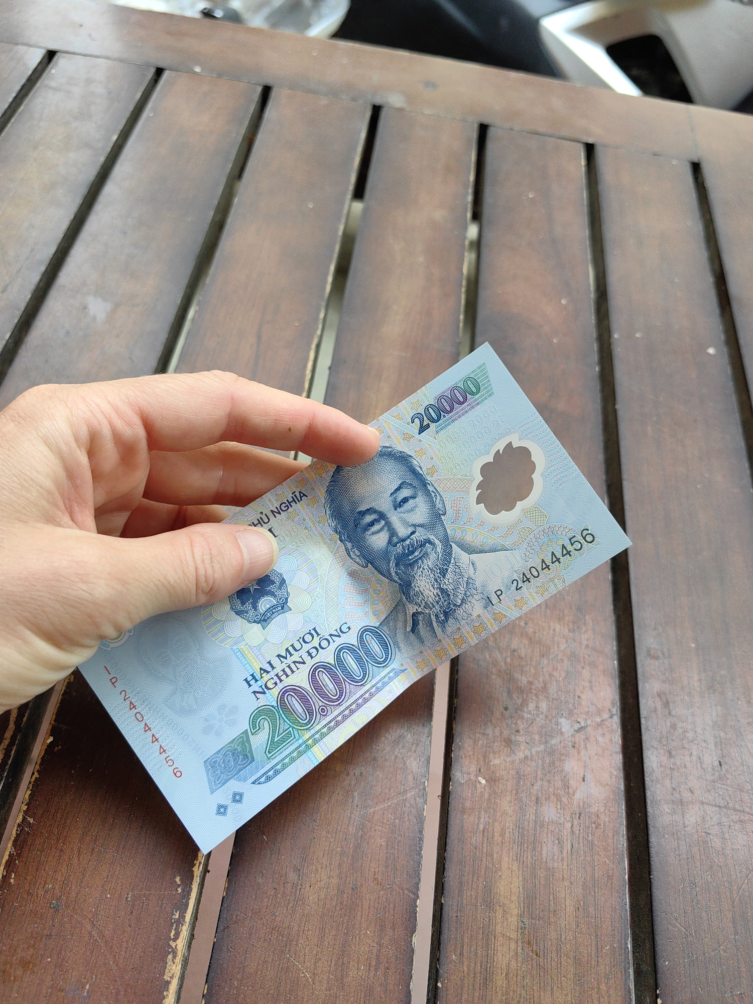 A human hand, appearing to be that of an adult, is holding a single 20,000 Vietnamese Dong (VND) banknote. The banknote is light blue, displaying the portrait of Ho Chi Minh on its front and the serial number IP 24044456. The hand grips the currency between its thumb and index finger. The scene is set against a worn, brown wooden slatted surface, likely a table, which exhibits visible grain and minor blemishes. Faint, out-of-focus background elements are visible above the wooden surface. The location is Sa Pa, Vietnam.