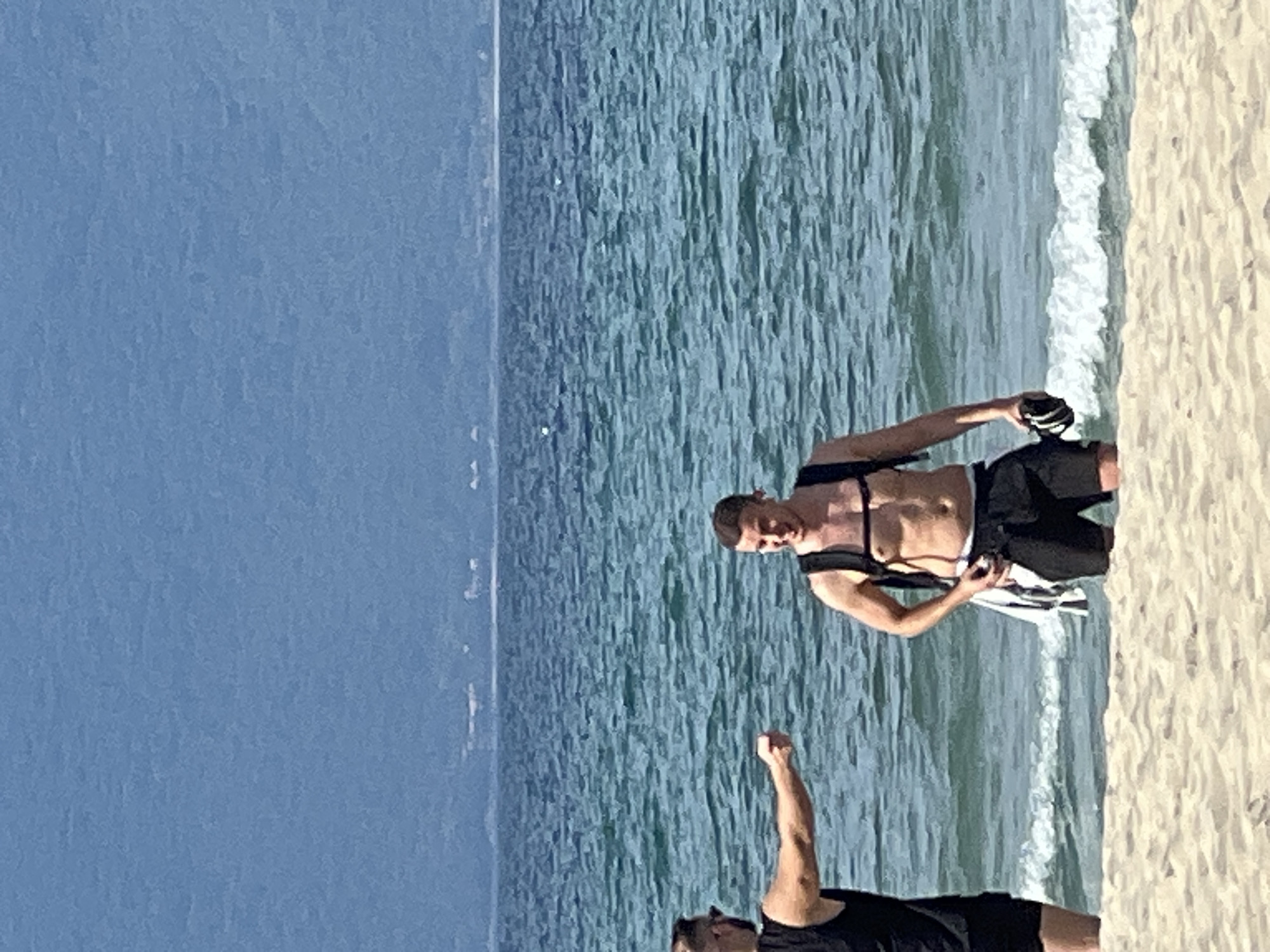 Two shirtless men are standing in the shallow water of a beach in Da Nang, Vietnam. The man on the left, wearing sunglasses and a black tank top, is pointing his right hand towards the man on the right. The man on the right is wearing black shorts and a backpack with a towel hanging from his waist. He is holding a pair of black shoes or sandals in his right hand and is smiling. The sea is a vibrant blue-green with small waves. In the background, a hazy blue mountain range can be seen in the distance, and the sky is a pale blue. The foreground consists of a sandy beach.