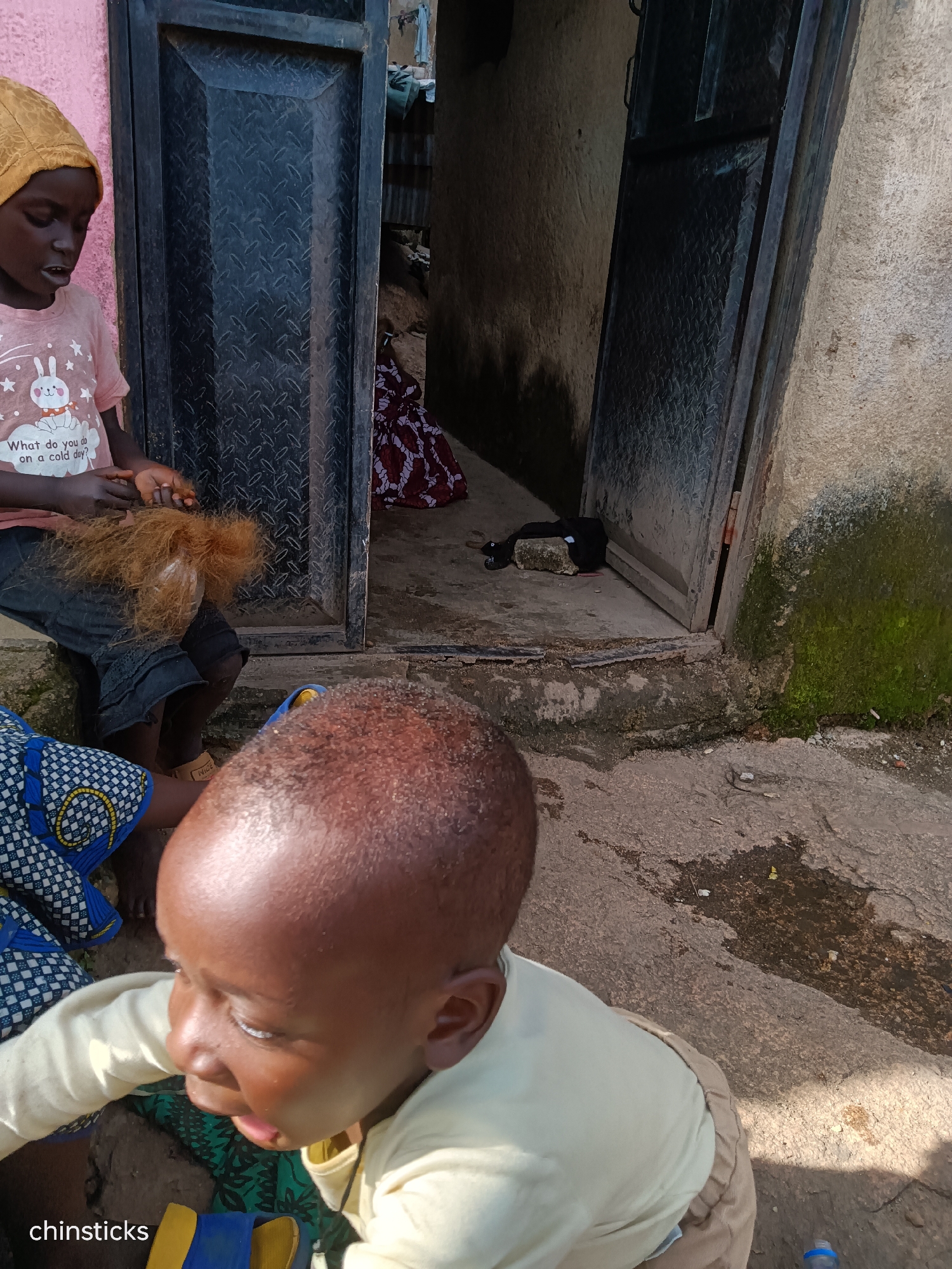 A young child, seen from the back, sits on a stone step outside a doorway. The child is wearing a light yellow long-sleeved shirt and tan pants. Their head is bald and shiny, and their right ear is visible. In front of this child, another child, likely older, is seated, facing to the left and slightly away from the camera. This child wears a pink t-shirt with a graphic of a rabbit and the text "What do you do on a cold day?". They are wearing dark shorts with lace trim. This child's hands are manipulating a bundle of reddish-brown, coarse material, possibly hair or fiber. Behind these children, an open doorway leads into a dark interior, where a person in a dark patterned garment is partially visible. The environment is an outdoor urban setting in Jos, Nigeria, characterized by rough concrete flooring, a textured metal door, and a rough, light-colored wall with patches of moss. A bottle cap is visible on the ground in the lower right corner.