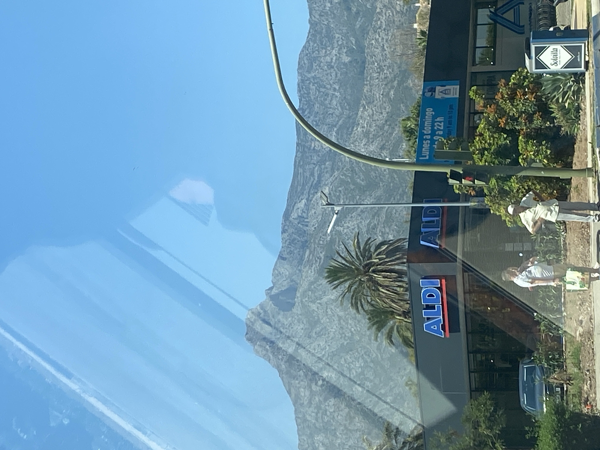 The image shows a reflection on a window. In the foreground, a blue sky is visible with some faint reflections of clouds or other objects. The main scene captured in the reflection is a street in Marbella, Spain. A modern building with the ALDI logo prominently displayed on its facade is visible. The building has large glass windows, and the reflection shows a street with a sidewalk, some greenery including palm trees, and a traffic light. Two people are seen on the sidewalk in front of the ALDI store. One person is wearing a light-colored shirt and pants, and the other is in a light-colored shirt and checkered shorts. A blue car is parked in front of the ALDI store. In the background, a large, rocky mountain range dominates the landscape. The sky is clear and blue, suggesting a sunny day. A sign on the building indicates opening hours: "Lunes a domingo 9 a 22 h," meaning Monday to Sunday from 9 AM to 10 PM. Another sign on the building has a blue "A" logo. A small white box with a diamond logo and the word "Sotavilla" is also visible on the side of the building. The lighting suggests it is daytime, possibly late morning or early afternoon due to the bright sunlight and clear sky. The overall scene depicts a commercial area in Marbella on a clear day.