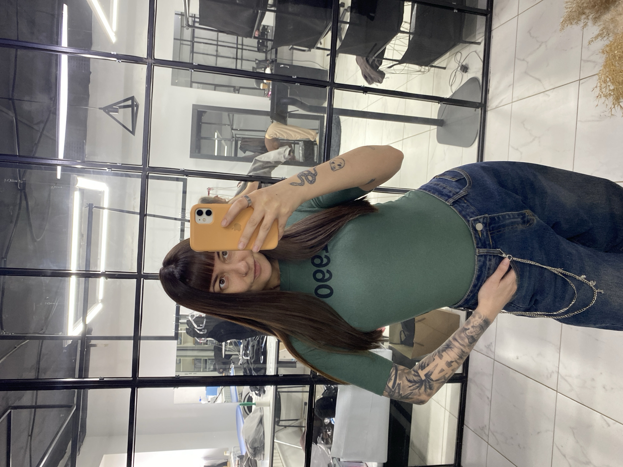 A woman with long dark hair and bangs is taking a selfie in a room with a grid-like window frame. She is holding an orange phone in her left hand. The woman is wearing a green shirt with black text on it and blue jeans with a silver chain. She has tattoos on both arms.
The room appears to be a salon, as evidenced by salon chairs and equipment reflected in the windows. The floor is white with a marbled pattern. The reflection in the window shows interior details including lighting, salon equipment, and the city of Votkinsk is not visible in the picture.