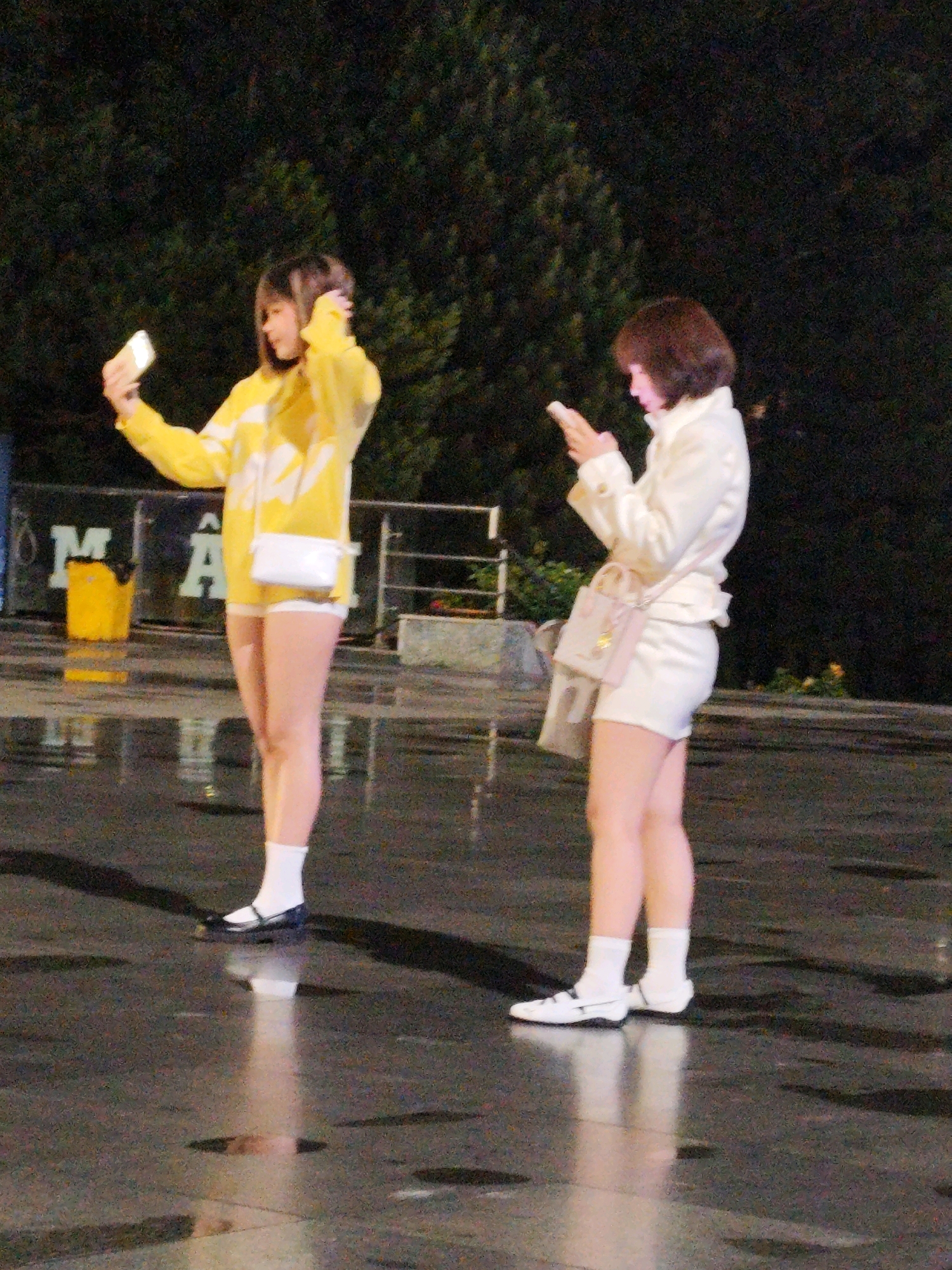 Two young women are standing on a wet, reflective surface in what appears to be a city square at night. The woman on the left is wearing a bright yellow hooded jacket with white stripes and white shorts. She has her hair down and is holding a white smartphone, taking a selfie. A small white purse hangs from her shoulder. The woman on the right is wearing a white jacket and white shorts. She has short, dark hair and is looking down at her white smartphone. She has a light pink purse slung over her shoulder, with a fabric shopping bag attached to it. Behind them, there are trees and a blurred background, suggesting an outdoor urban setting. In the background, on the left, a yellow trash bin is visible next to a sign with the letters "M" and "A". The ground is slick and wet, reflecting the lights from the surrounding area. The overall scene suggests a casual night out in Da Lat, Vietnam, possibly near a tourist area or a lively public space.