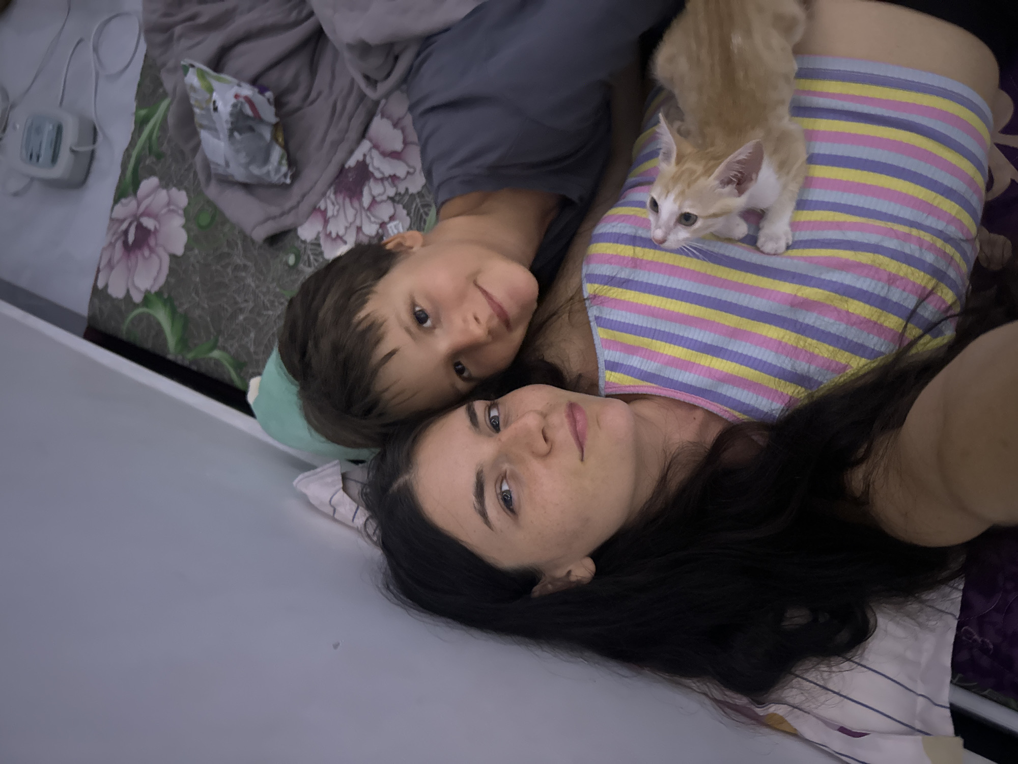 In this overhead, slightly angled shot, a young child and an adult, likely mother and son, lie in bed with a small ginger kitten. The child, with short brown hair and bright eyes, smiles at the camera, his head resting on the adult's shoulder. The adult, with long dark hair and freckled skin, looks up and to the side, a gentle expression on her face. The kitten, perched on the adult's colorful striped top, gazes curiously to the left. The background shows a bed with floral-patterned sheets and a gray blanket, and to the left, a small electronic device is visible. The scene appears intimate and relaxed, suggesting a cozy moment at home, possibly in the morning given the soft, diffused lighting. The presence of a kitten and the casual attire hint at a peaceful, domestic setting. The location context provided is Mũi Né, Vietnam, though there are no specific visual cues in the image to confirm this.
