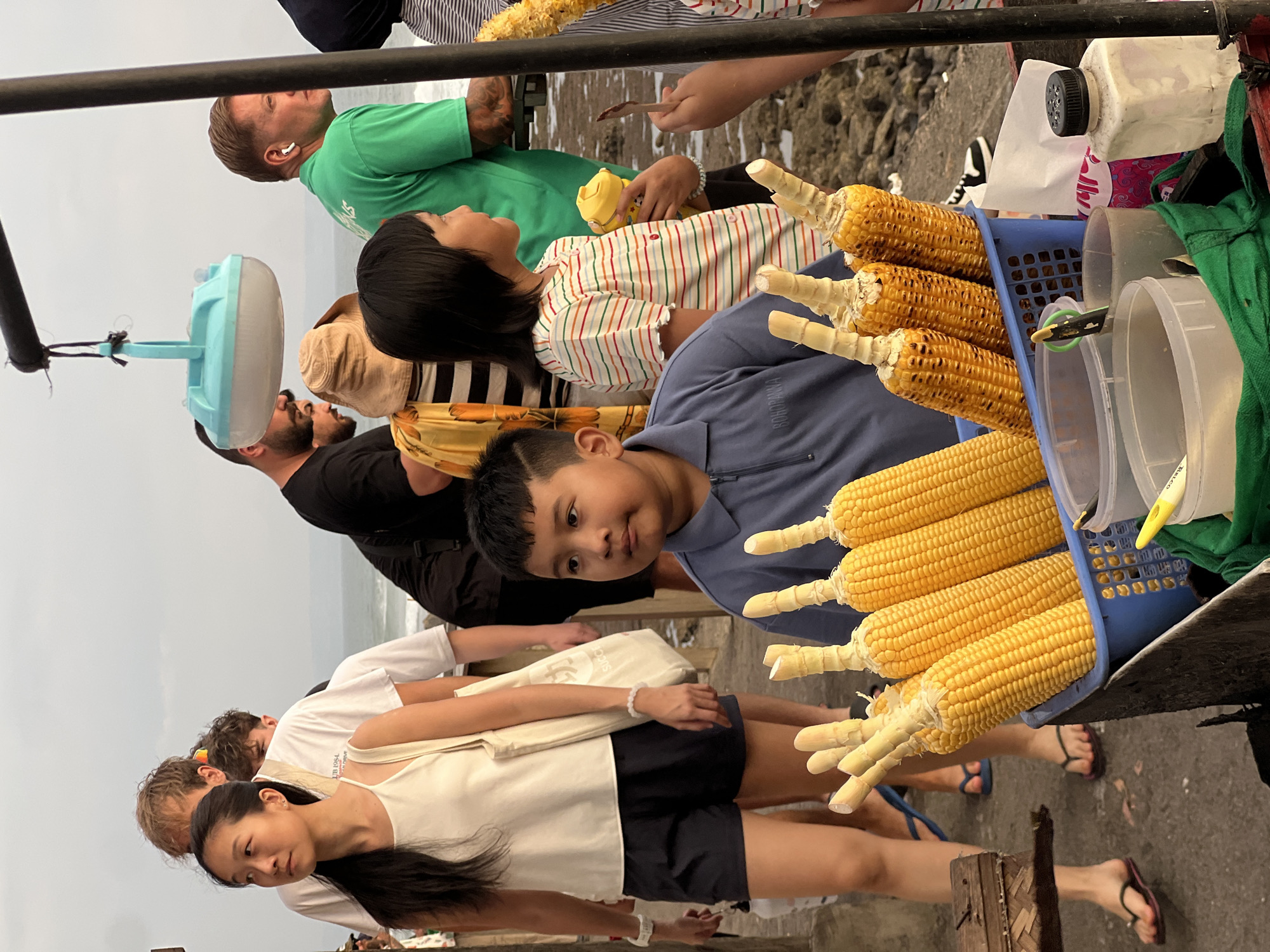 In North Kuta, Indonesia, on a paved area near the ocean, a boy in a blue collared shirt is standing next to a cart selling corn. He is looking to his left. Several cobs of corn are arranged in a blue plastic basket in front of him, some with char marks from grilling. Behind the boy, a group of people are walking, including a woman in a white tank top and dark shorts, and a man in a green shirt wearing earbuds. A light fixture hangs from a pole above the cart. The sky is overcast.