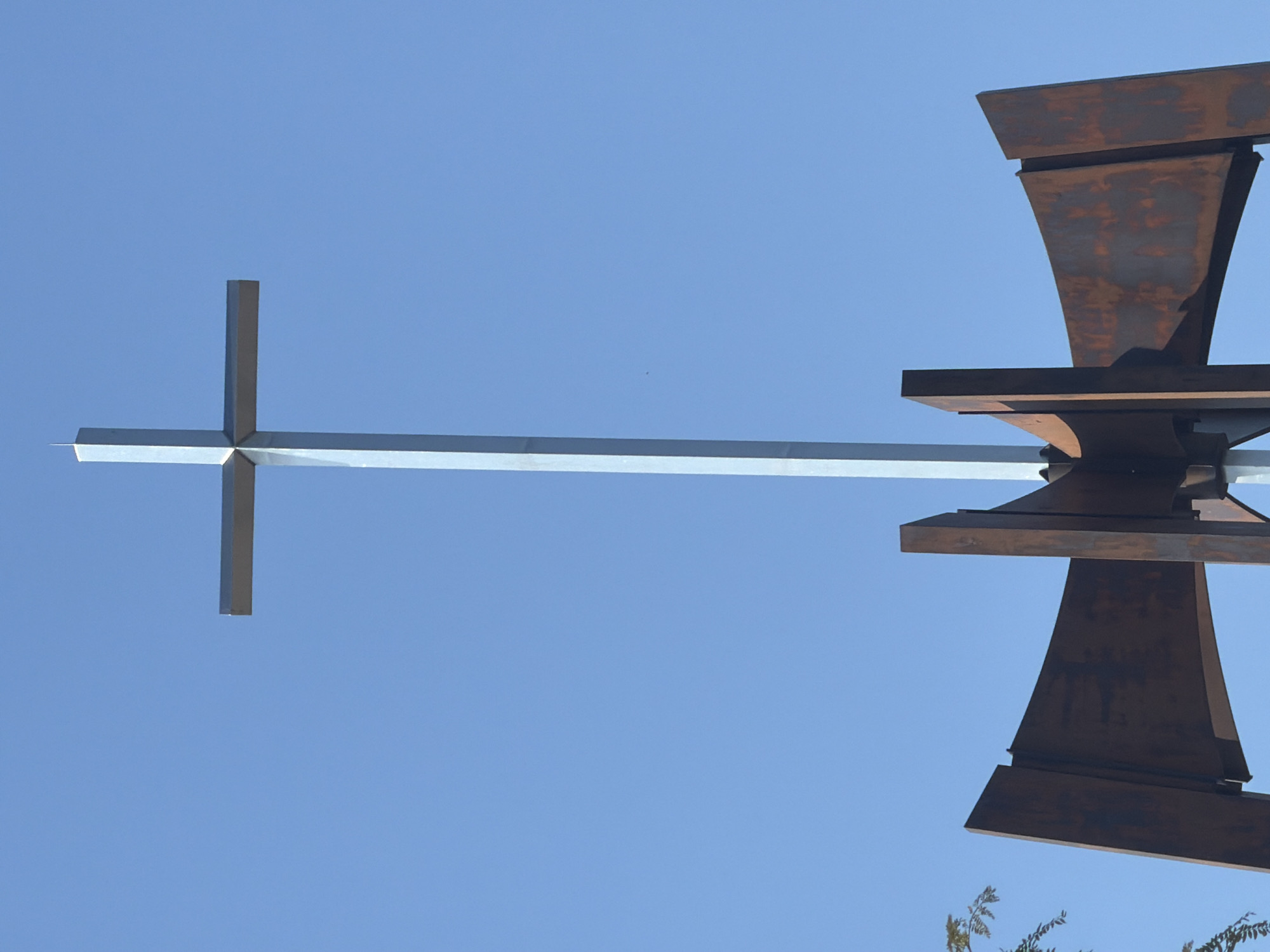 The photograph features a metal sculpture set against a clear, cloudless blue sky. Part of the sculpture consists of a long, slender silver cross or plus sign shape extending horizontally across the frame. To the right, there are several overlapping metal pieces that appear to be rusted or have a patina, creating a contrast in color and texture. The overall design of this segment resembles a stylized wheel or turbine. Hints of foliage are visible at the bottom right corner of the frame. The location is identified as Niles, United States of America.