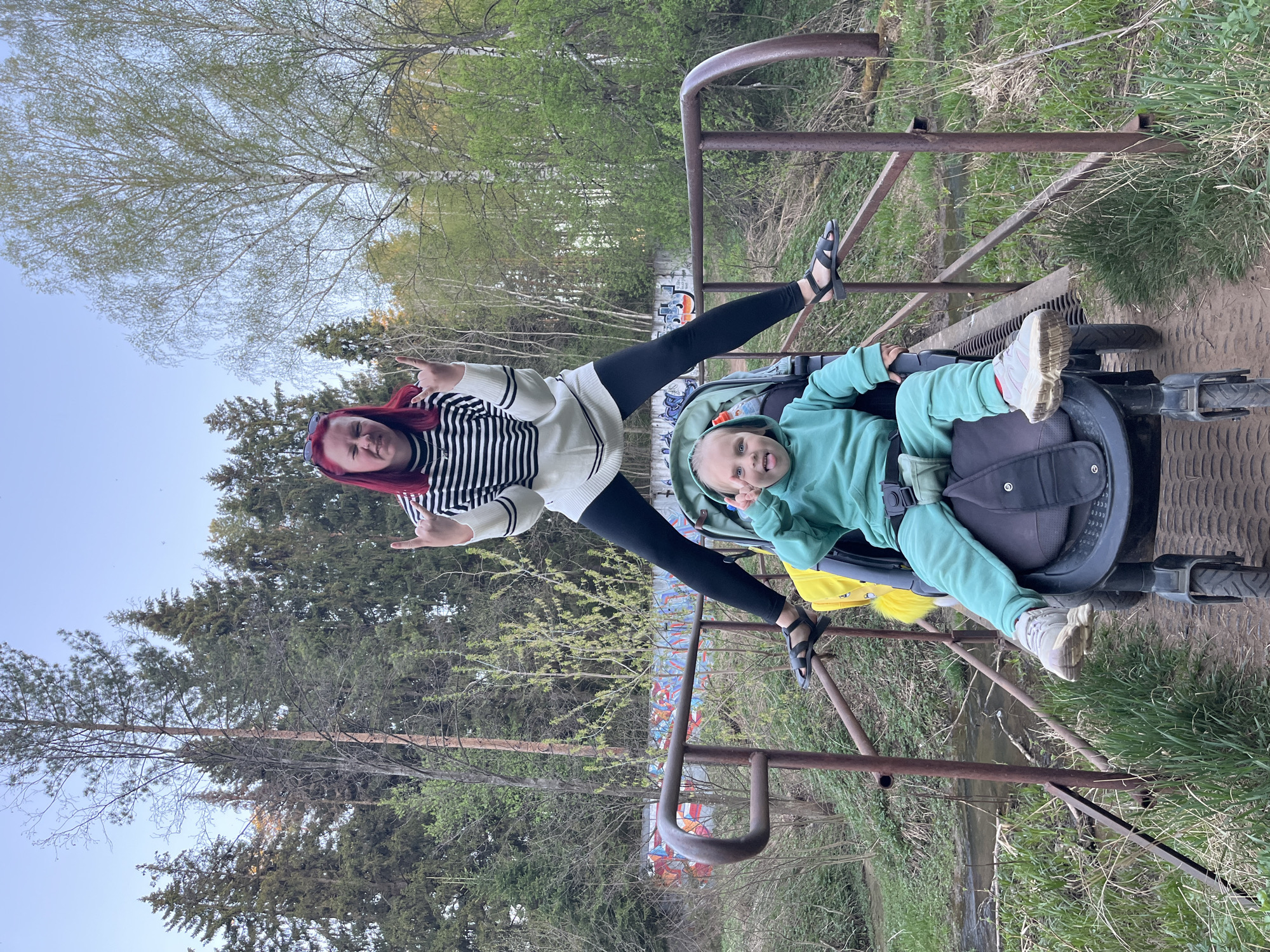 A woman with red hair and a child are posing on a rusty metal structure. The woman, wearing black leggings and a black and white striped top, is making a hand gesture. The child, dressed in a mint green outfit, is in a stroller. The background features a forest and some graffiti on a wall. The picture was taken in the city of Chaikovsky, Russia.