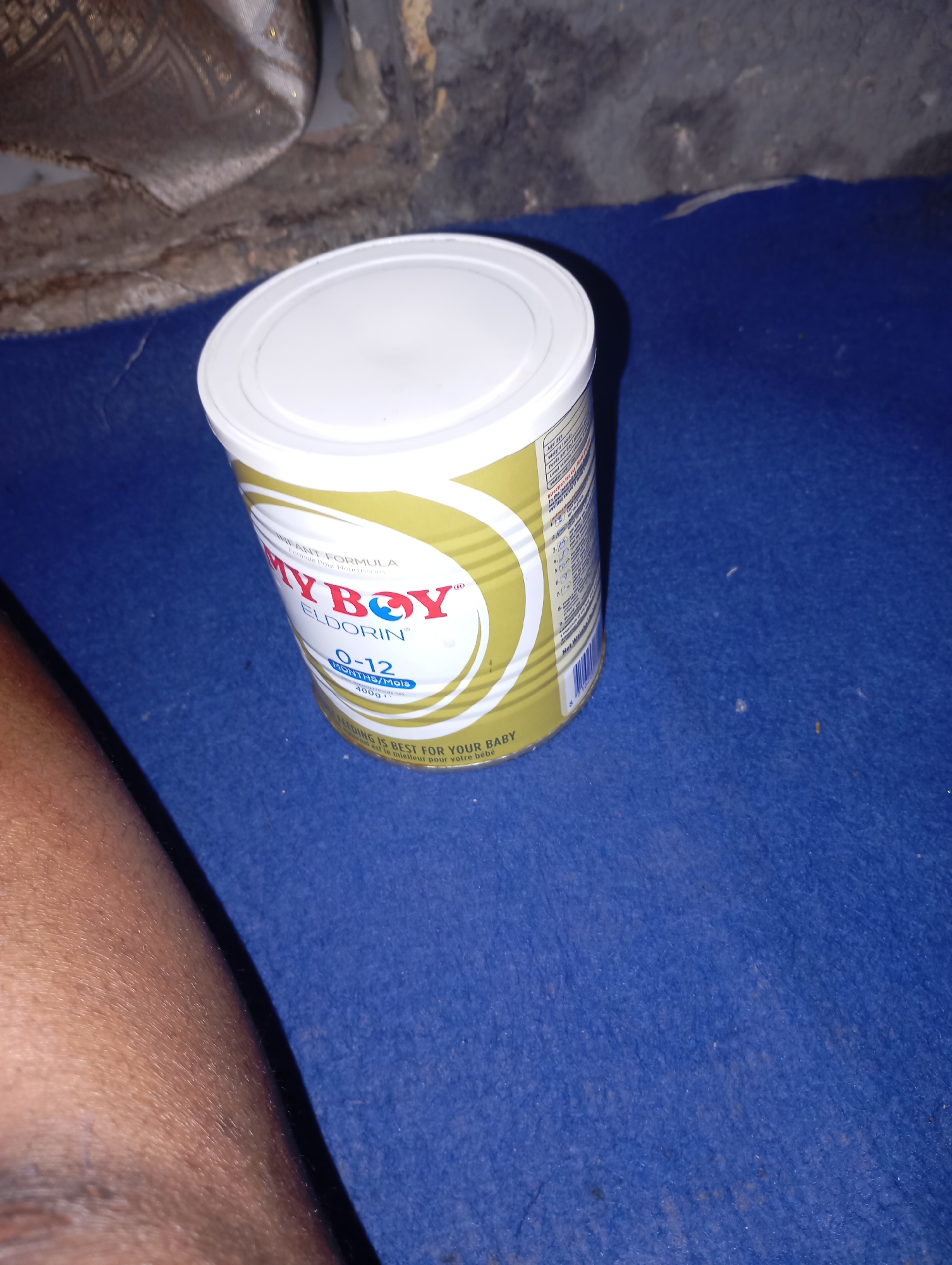 A close-up, eye-level shot shows a can of "MY BOY" infant formula on a blue fuzzy surface. The can has a white lid and a gold and white label with text in English and French. The label prominently features the brand name "MY BOY" in red letters. Below the brand name, it states "ELDORIN" and "0-12 MONTHS/Mois". Further text indicates "400g" and "INFANT FORMULA". The bottom of the label reads "FEEDING IS BEST FOR YOUR BABY" in English and "L'ALIMENTATION EST LE MEILLEUR POUR VOTRE BÉBÉ" in French. To the left of the can, a person's leg, with visible skin texture and hair, is partially visible. The background consists of a dark, textured wall and a brown, patterned fabric. The lighting is dim, casting shadows. The setting appears to be indoors, possibly a residential area.