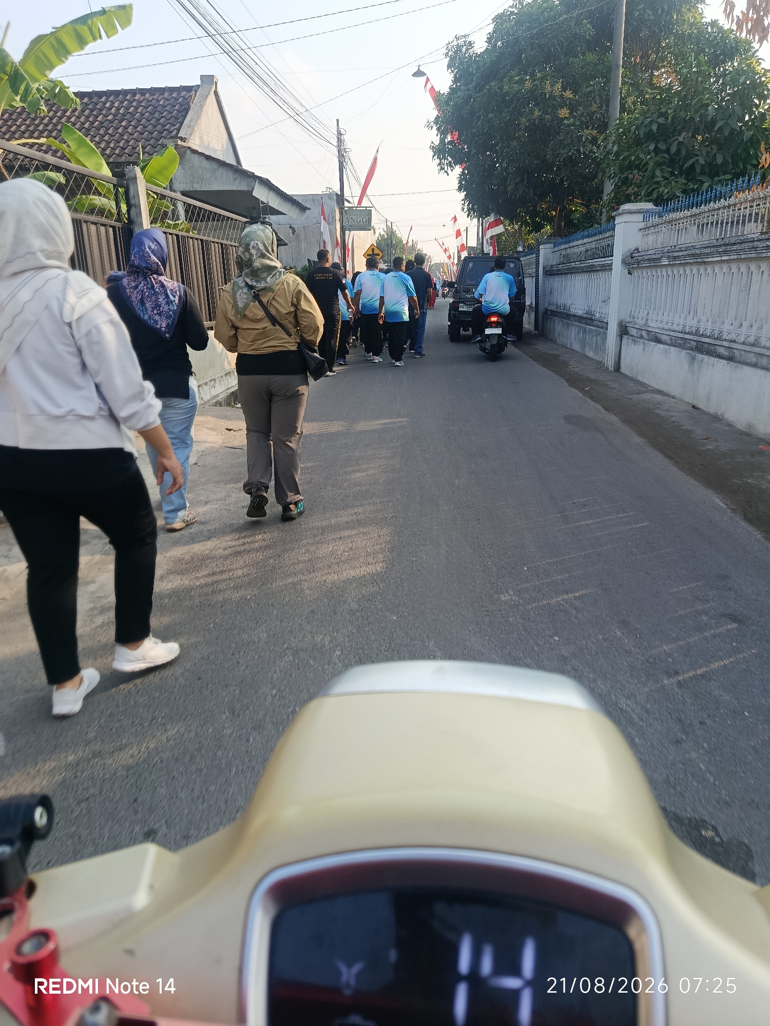 In Balerejo, Indonesia, a group of people are walking down a paved road lined with fences and houses. The people are walking in the same direction, and some are wearing headscarves. One person is riding a motorcycle. The scene appears to be a street during the day, with a clear sky and the sun shining. The image also shows the timestamp "21/08/2026 07:25" and "REDMI Note 14" in the lower part of the image.