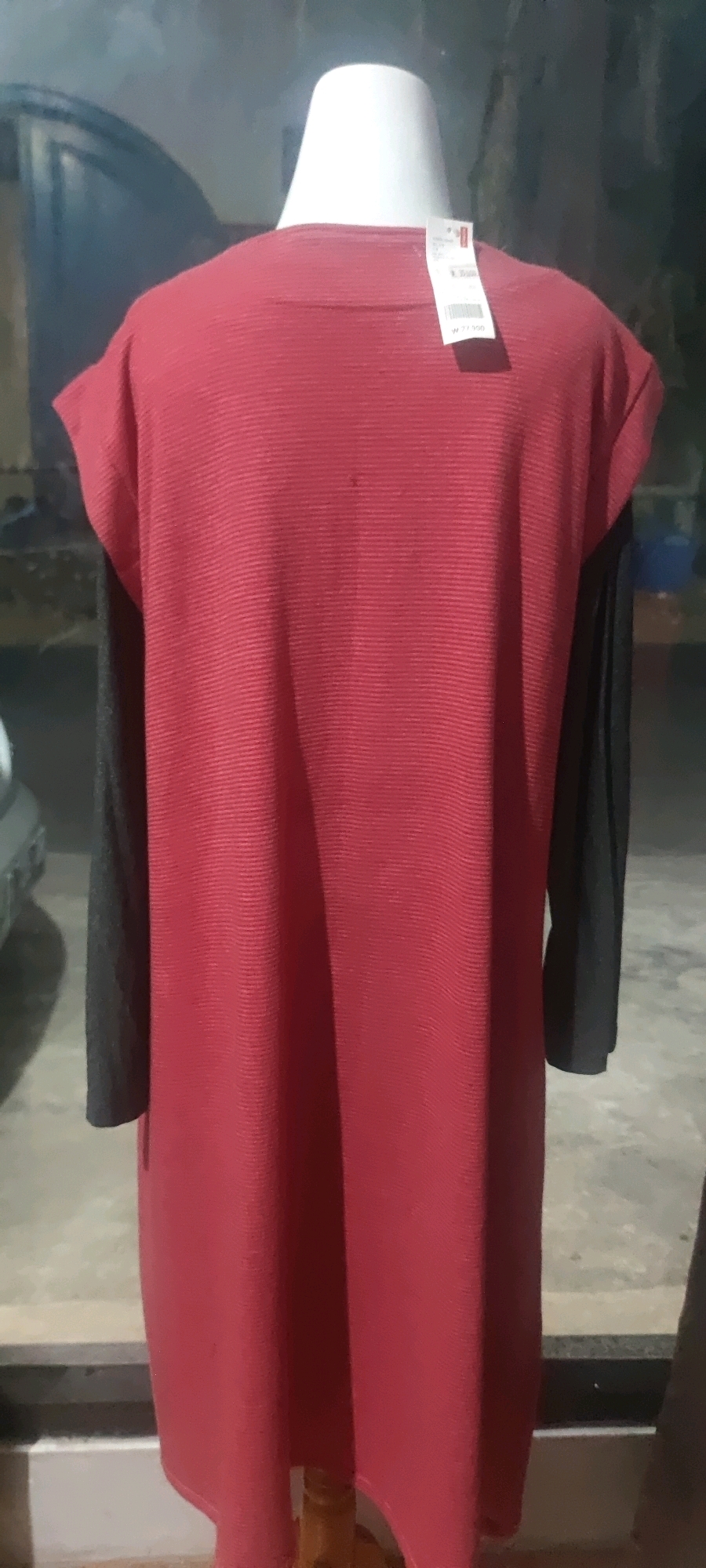 A red dress with black sleeves is displayed on a white mannequin. A tag is attached to the back of the dress near the neckline. The mannequin is positioned indoors, with a blurred background that appears to be a reflection in a window. The environment suggests a retail setting.