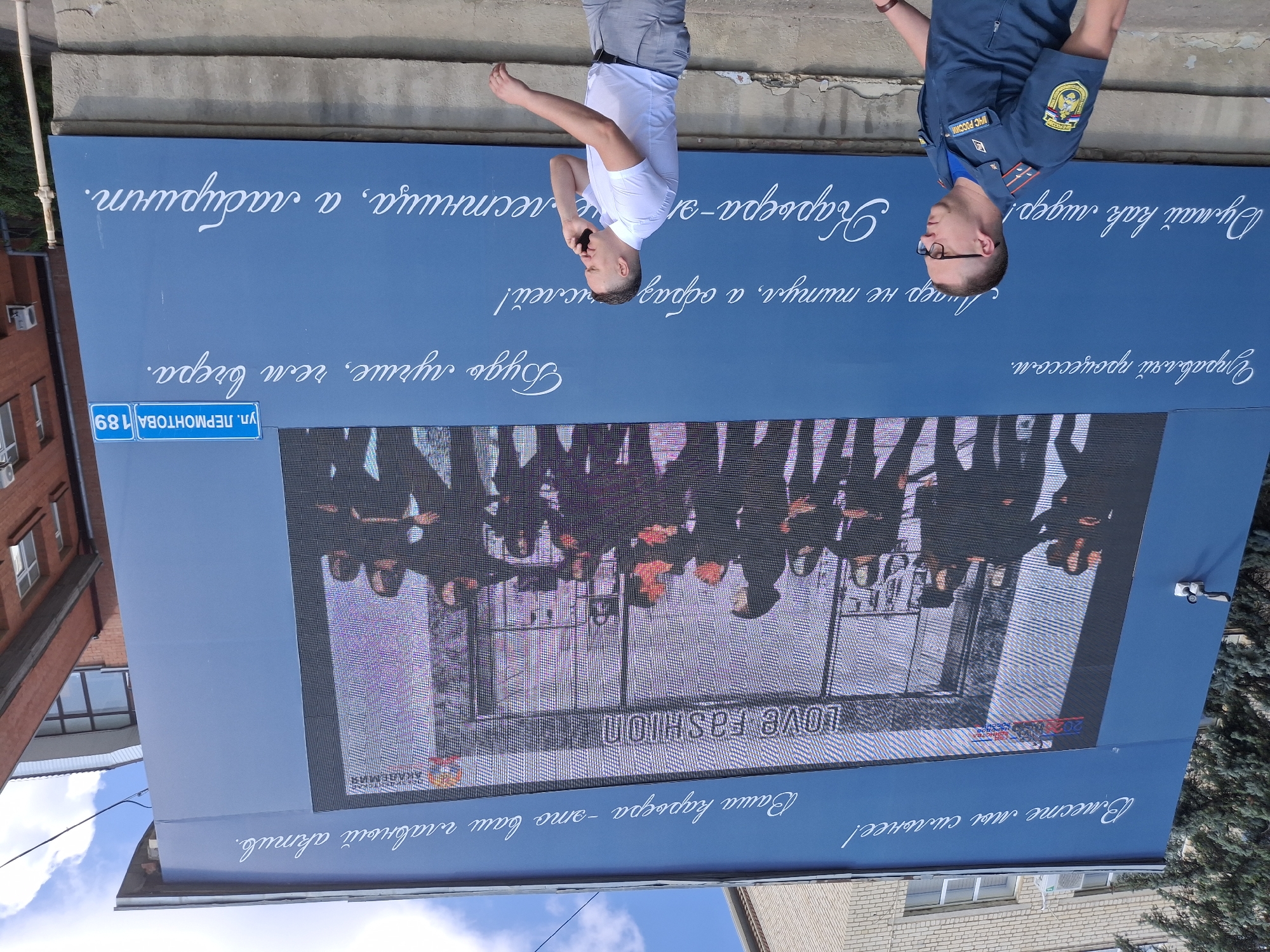 The image shows a large blue billboard on the side of a building, displaying text in white cursive font. The billboard features a digital screen showing a fashion-related advertisement with the text "LOVE FASHION". Two men are walking in front of the billboard. The man on the left is wearing a dark blue uniform with patches that read "МЧС РОССИИ" and a circular emblem, indicating he is a member of the Russian Ministry of Emergency Situations. He has short hair and glasses. The man on the right is wearing a white short-sleeved shirt and grey pants, and is talking on a mobile phone. He has short hair. In the upper right corner of the billboard, a blue street sign is visible that reads "ул. ЛЕРМОНТОВА 189", which indicates the location as Lermontova Street, house number 189, in Stavropol, Russia. The background shows parts of buildings and a blue sky with clouds.
