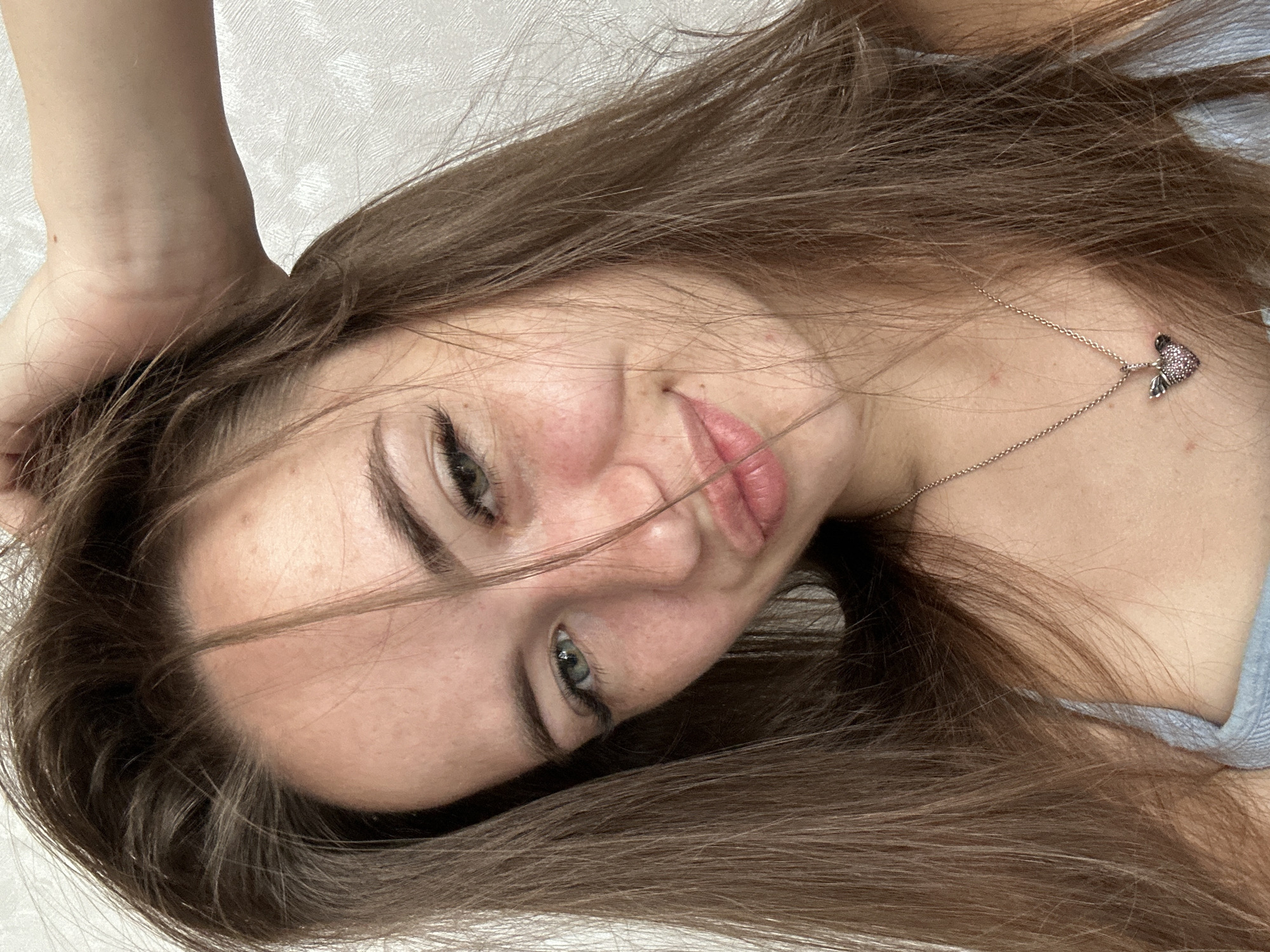 The image is a close-up selfie of a young woman with long, brown hair. She is looking at the camera and has her hand on her head. Her skin has freckles, and she is wearing a light blue top and a necklace with a heart-shaped pendant. The background is a plain, textured wall. The scene appears to be indoors.