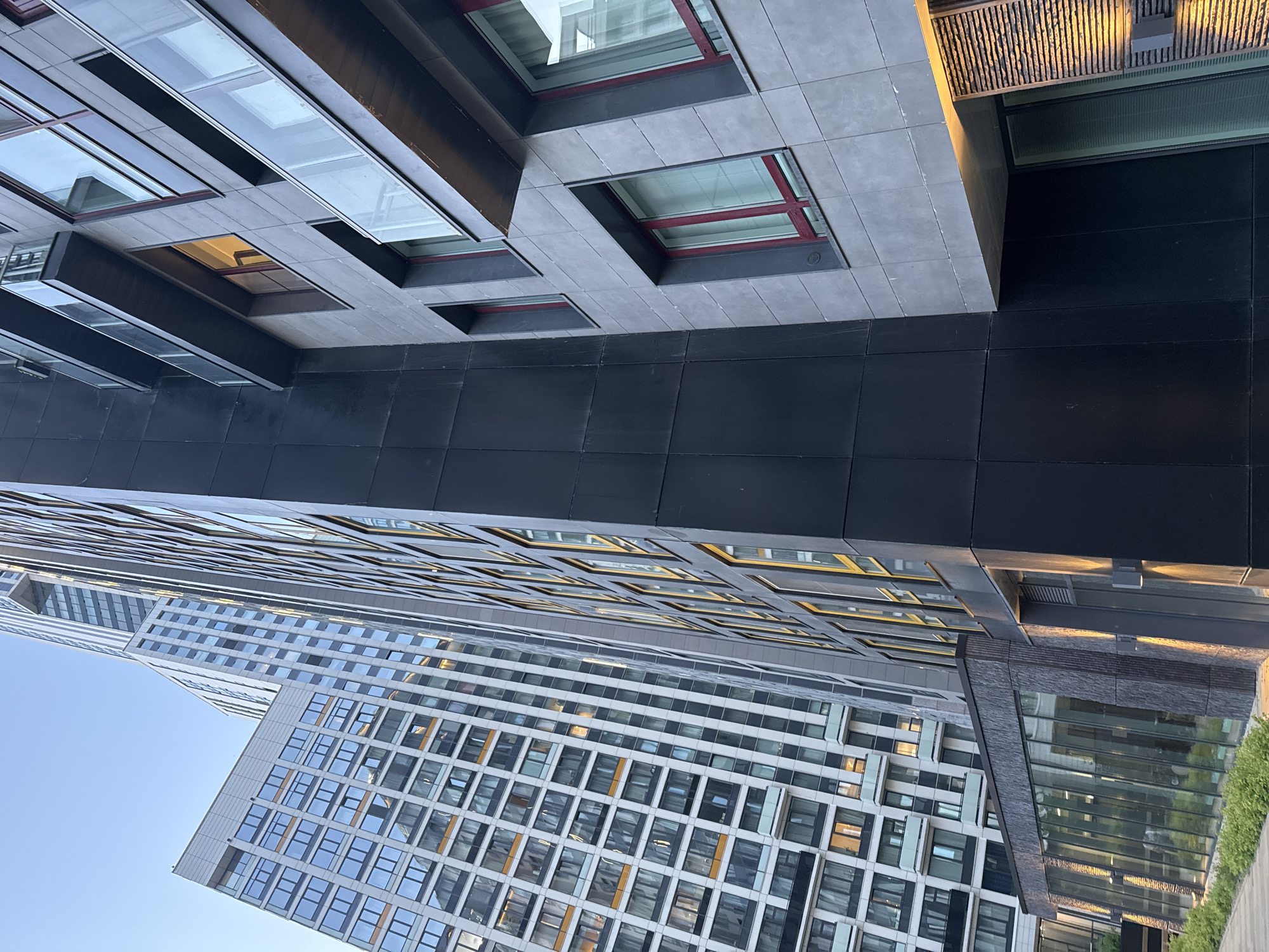 The image presents a modern architectural scene in Khimki, Russia, captured from a low angle looking upwards towards multiple tall buildings under a clear blue sky.
In the foreground, the lower section of a building features a dark grey or black paneled facade. Above this, a lighter grey tiled structure displays various rectangular windows, some with distinct red frames, and integrated glass skylights. One window emits a warm, yellowish light from within. Adjacent to this, a textured architectural element, possibly a canopy or decorative screen made of slats, is illuminated by a warm, golden light, creating a strong contrast.
Rising prominently in the midground and background are several contemporary high-rise buildings. Their facades are characterized by a repetitive grid pattern of numerous square and rectangular windows set into light-colored, likely grey or white, panels. Many windows reflect the blue sky and surrounding structures, while others reveal interior lights. The clean lines and geometric design emphasize the modern aesthetic of the urban environment.
To the right side of the image, near the base of one of the buildings, a section of vibrant green foliage is visible, possibly part of a landscaped area or rooftop garden. Large glass panels nearby reflect this greenery.
The lighting suggests either late afternoon or early evening, with warm sunlight or artificial illumination accentuating certain architectural details. The weather appears clear with no clouds. No people or visible text are present in the image. The overall impression is one of sleek, functional, and contemporary urban development.
