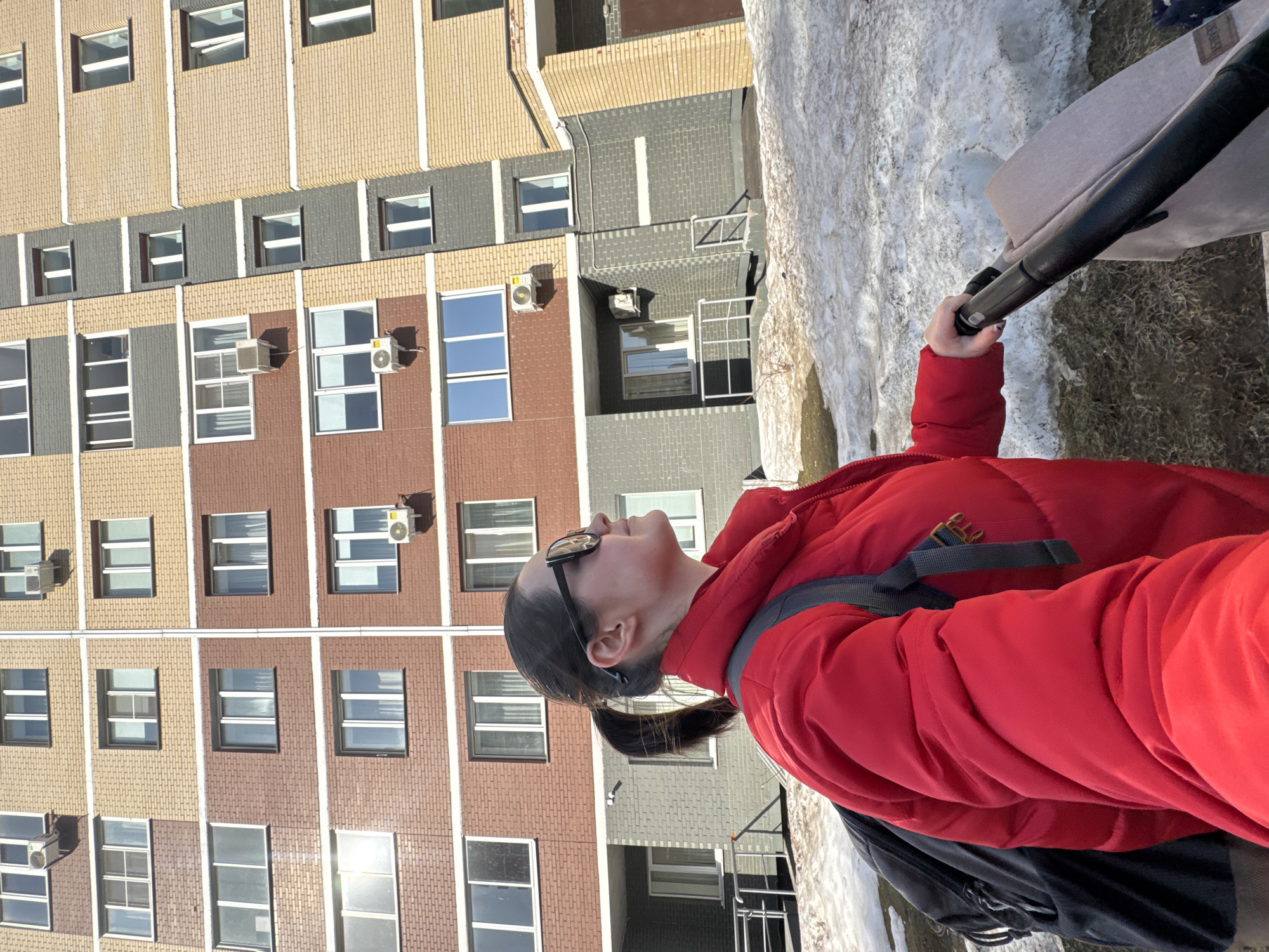 A woman in a red puffer jacket, sunglasses, and with a ponytail is standing outside, holding onto the handle of a stroller. She is looking up and to the left. Behind her is a multi-story building with beige, brown, and gray brick sections, and numerous windows, some with air conditioning units. Patches of melting snow are visible on the ground. The location is Izhevsk, Russia.
