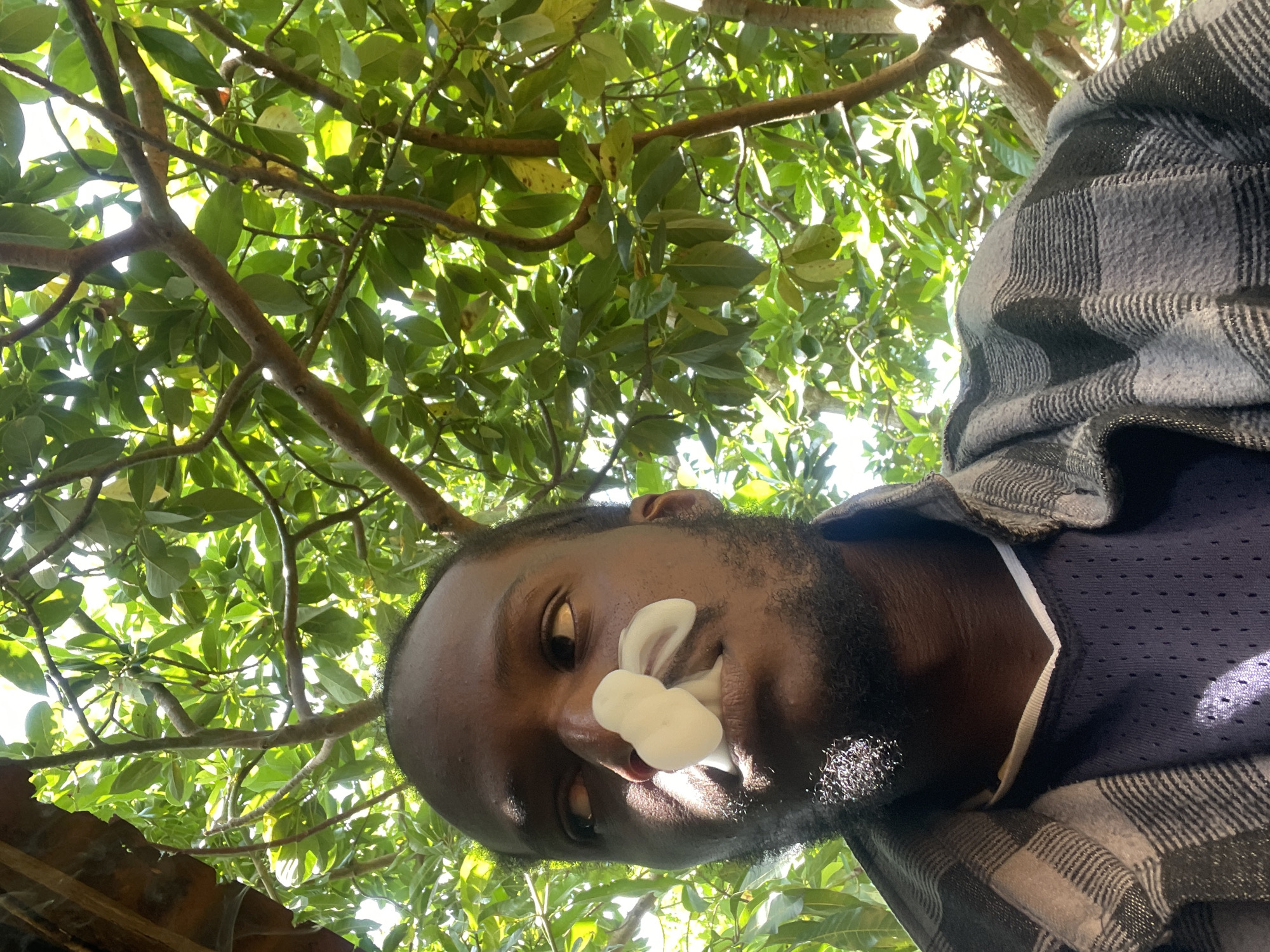 A close-up, selfie-style shot captures a Black male subject from the chest up, looking to his left, with white smoke emanating from his mouth and nose, creating a cloud-like effect. The subject has dark skin, a short beard, and is wearing a dark blue athletic jersey underneath a grey and black plaid shirt. He is outdoors, with lush green foliage and branches from trees filling the background, suggesting a natural, possibly shaded environment. The lighting indicates it is daytime, with sunlight filtering through the leaves. The background appears to be consistent with the natural vegetation found in areas like the generally wooded and vegetated outskirts of Jos, Nigeria, where such outdoor settings are common.