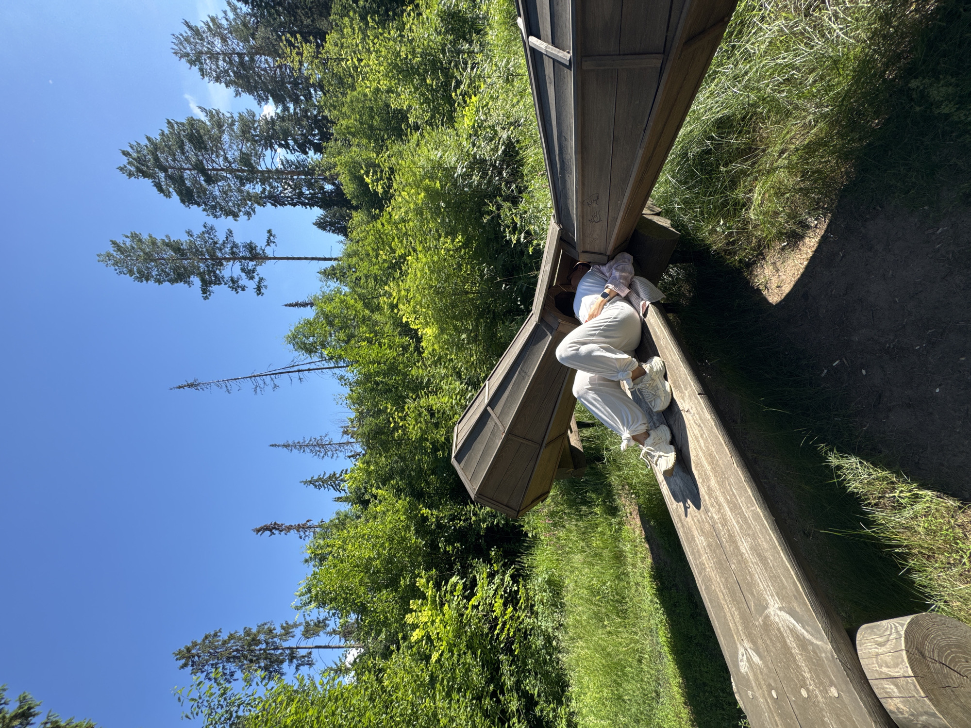 The image shows a person lying down on a wooden structure in a forest. The person is wearing white pants and a white shirt. The wooden structure appears to be a large, elongated object with a curved shape, possibly part of an art installation or playground equipment. The surrounding environment is a dense forest with lush green trees and a clear blue sky. The ground is covered with grass and dirt. The lighting suggests it is daytime with bright sunlight. The location is identified as Votkinsk, Russia.