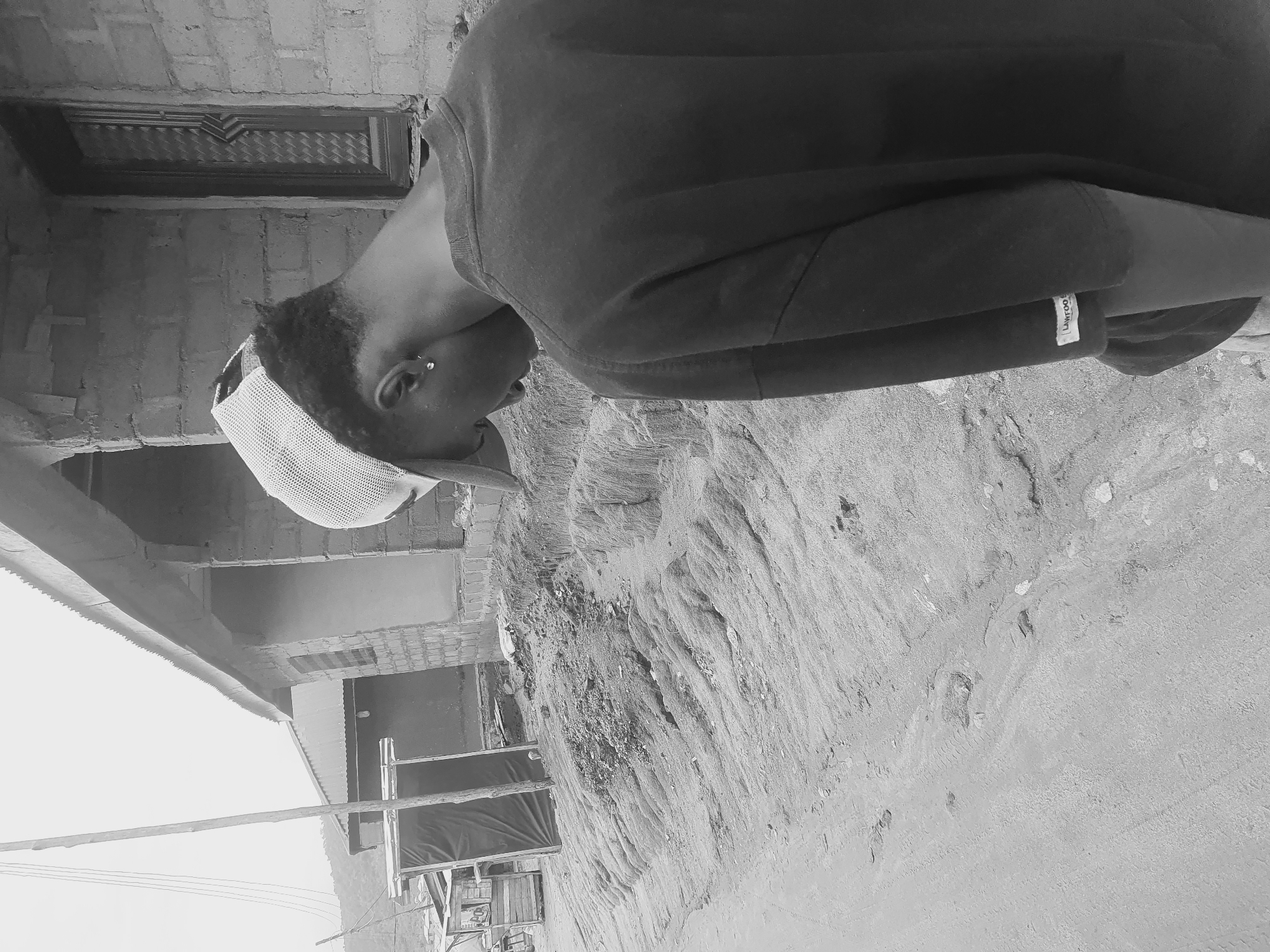 A grayscale medium shot depicts a person with dark hair wearing a baseball cap, a dark long-sleeved shirt, and an earring. The person is looking down and facing left, in the city of Lokoja, Nigeria. A portion of a building is visible to the left. The ground is rough and sandy. A partially visible structure is in the background to the left.
