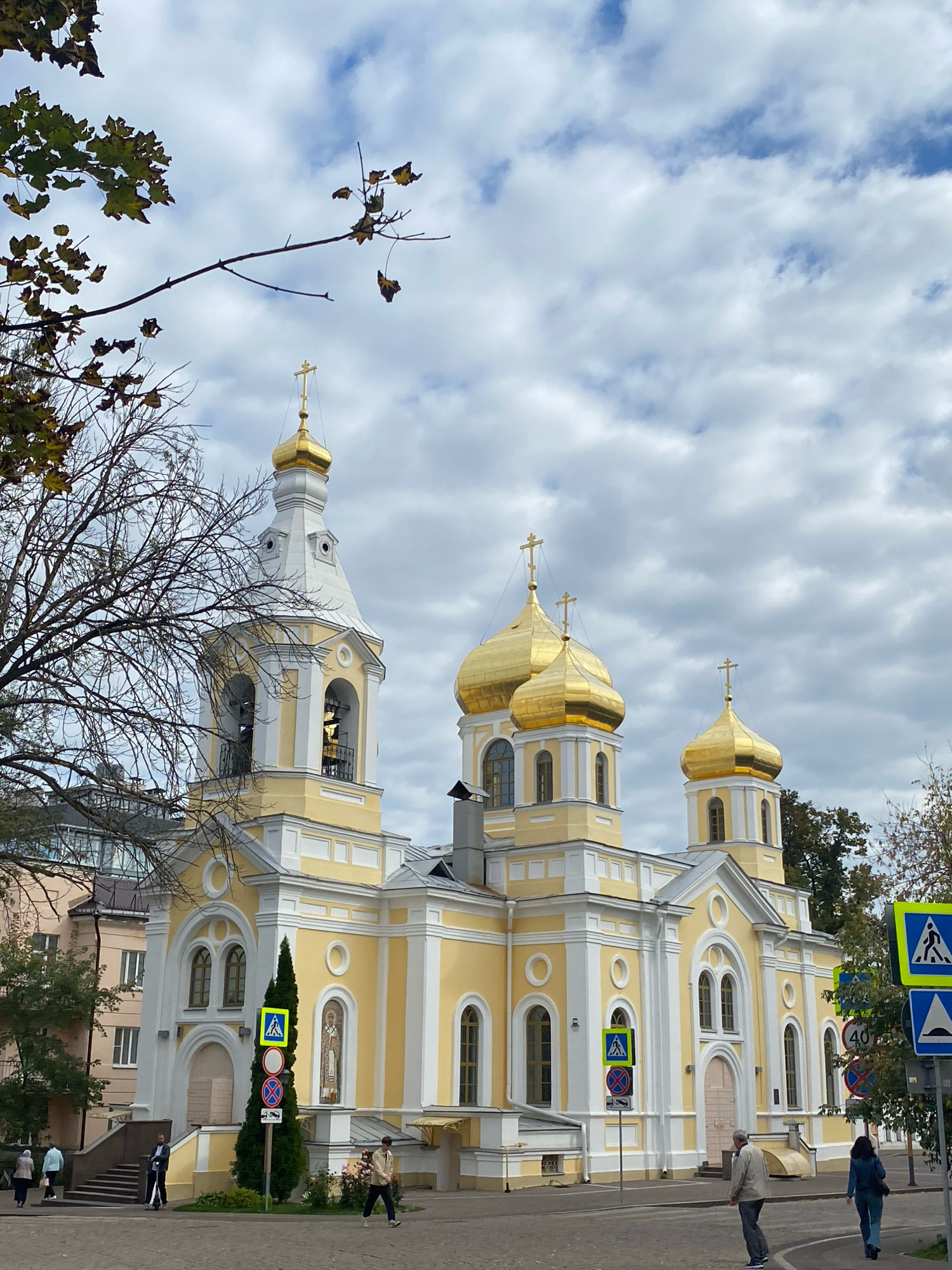 A bright yellow and white Eastern Orthodox church with golden domes stands prominently against a cloudy sky. A tall bell tower with a single bell is visible to the left of the main structure. Several golden domes with crosses adorn the church's roofline. The church is situated in an urban environment, with a street and sidewalk in the foreground. Various traffic signs, including pedestrian crossing signs and speed limit signs, are present. A few people are walking on the sidewalk and crossing the street. Trees with autumn foliage are partially visible on the left side of the frame. The overall scene suggests an overcast day in the city of Nizhny Novgorod, Russia.