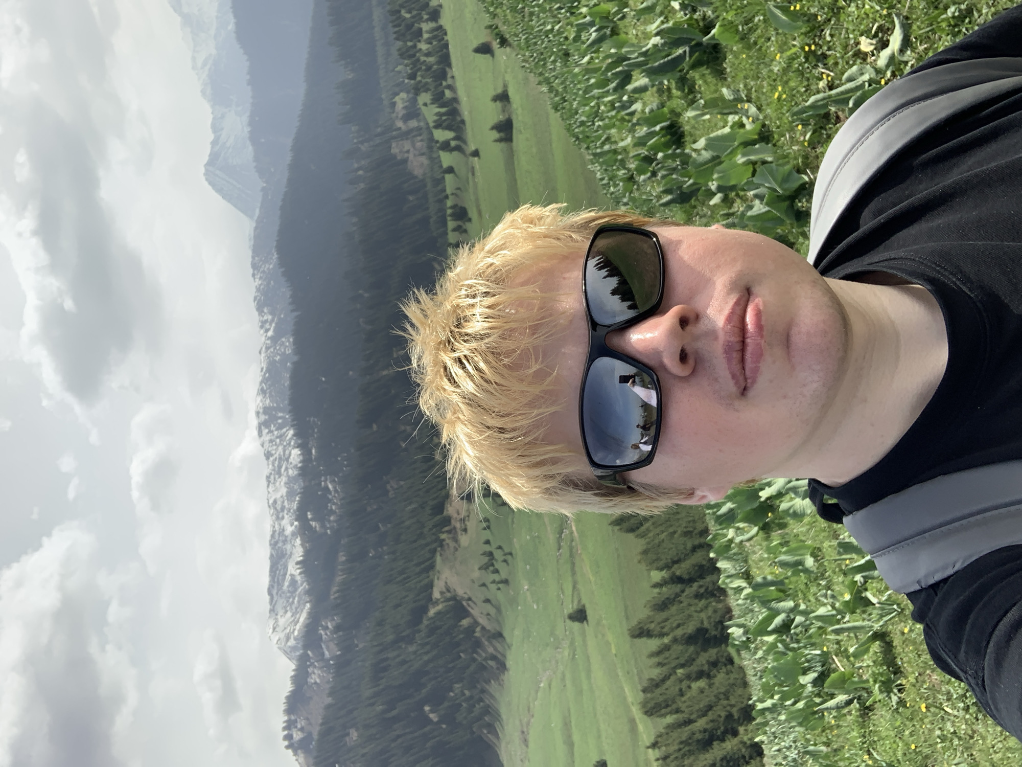A selfie of a blonde-haired person wearing sunglasses with a reflection of other people in the lens. The background features a mountain range with low-hanging clouds and a green valley. The photograph was likely taken near Almaty, Kazakhstan, given the local terrain. The person is wearing a black shirt with a gray backpack strap visible. The ground near the person is covered with green plants.