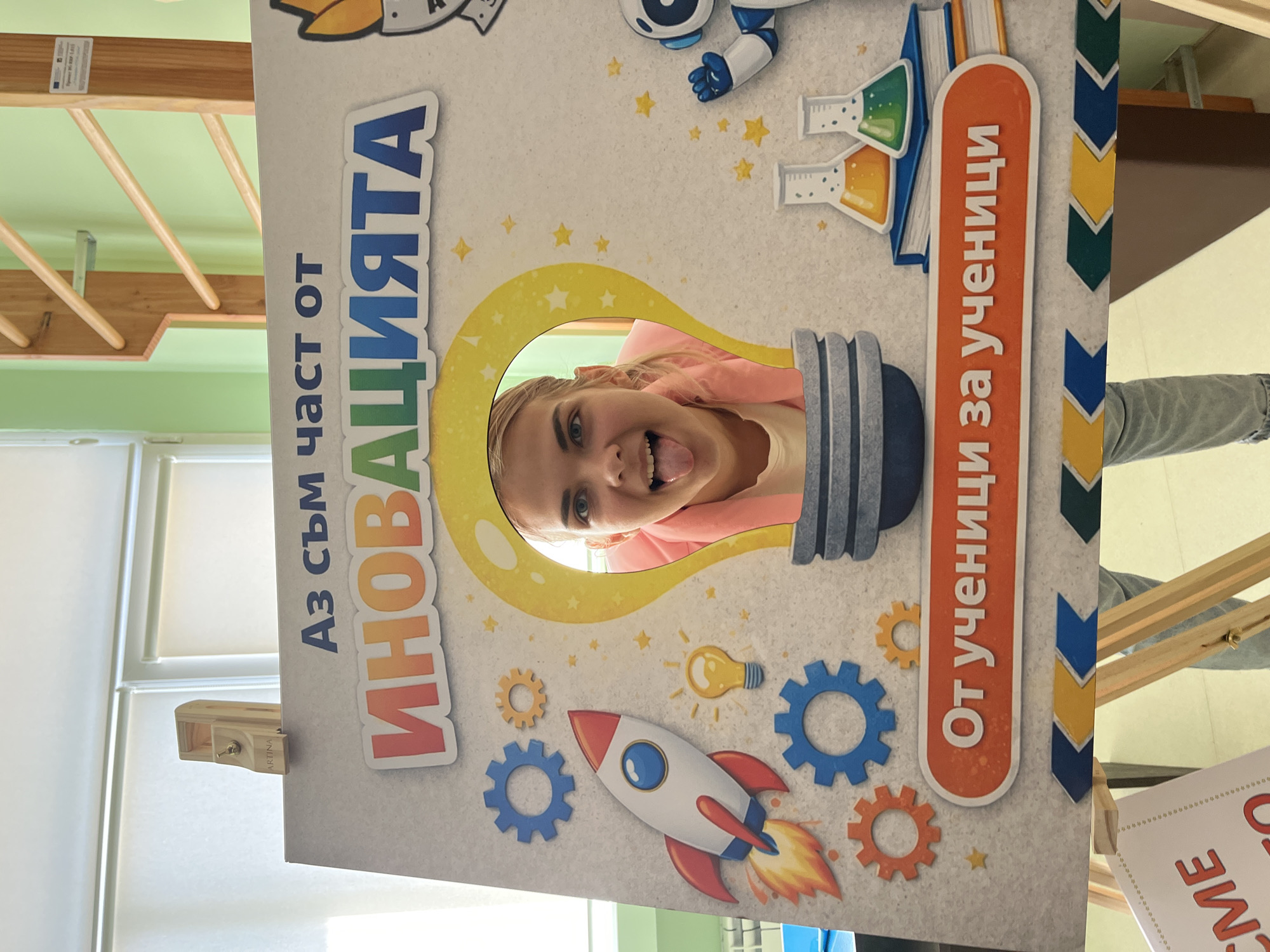 The image depicts a close-up, eye-level shot of a playful photo booth prop with a young woman's head peeking through a lightbulb cutout. The prop is predominantly white with colorful illustrations of a rocket, gears, lightbulbs, and beakers. The text on the prop, in Bulgarian, reads "Аз съм част от ИНОВАЦИЯТА" (I am part of INNOVATION) and "От ученици за ученици" (From students for students).
The woman, with blonde hair, is sticking out her tongue and smiling at the camera. She is wearing a pink cardigan over a white top. The prop is supported by an easel, visible at the bottom of the frame. In the background, there are hints of a classroom or school environment, including windows with blinds, a wooden climbing frame with colorful bars, and a light green wall.
A sign partially visible at the bottom left reads "CME". The overall scene suggests an event or activity related to innovation and student engagement, likely at a school in Zlatograd, Bulgaria, given the context provided.