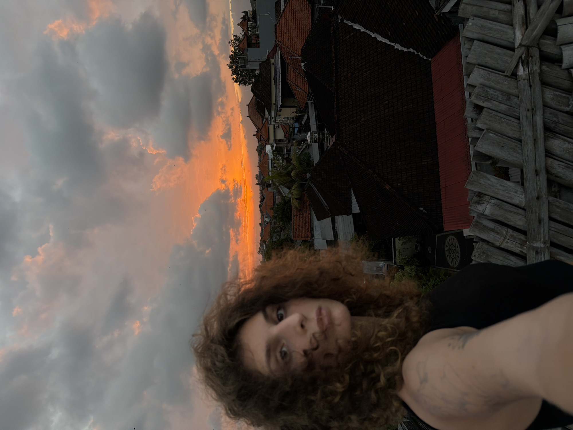 This media file depicts a selfie taken by a young woman with curly brown hair during sunset in North Kuta, Indonesia. The sky is a dramatic mix of dark clouds and vibrant orange and pink hues from the setting sun, creating a beautiful backdrop.
The woman is positioned in the foreground, with her face and upper body visible. She is looking directly at the camera with a playful expression, her lips slightly puckered. Her curly hair frames her face, and a tattoo is visible on her right arm. She is wearing a black top.
In the background, a cluster of buildings with traditional terracotta tiled roofs is visible, interspersed with greenery. The architecture suggests a tropical location. To the right of the frame, there is a weathered wooden structure, possibly part of a rooftop or balcony, adding a rustic element to the scene.
The overall impression is one of a casual, spontaneous moment captured during a picturesque sunset. The combination of the dramatic sky, the woman's engaging gaze, and the local architecture creates a sense of place and atmosphere. The time of day is clearly sunset due to the sky's colors. The location is identified as North Kuta, Indonesia, further enriching the context. No text is visible in the media file.
