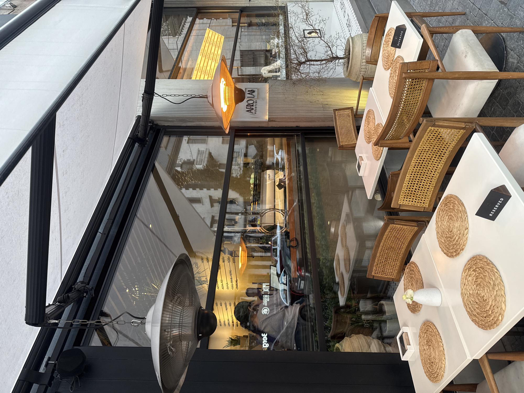 The image shows an outdoor seating area of Afro Lab Roastery, Yerevan, Armenia. The tables are set with placemats and reserved signs. Wicker chairs surround the white tables. A fan is mounted on a wall next to the dining area. A sign for the roastery is visible with a hanging lamp. The reflection in the window shows people inside the coffee shop.