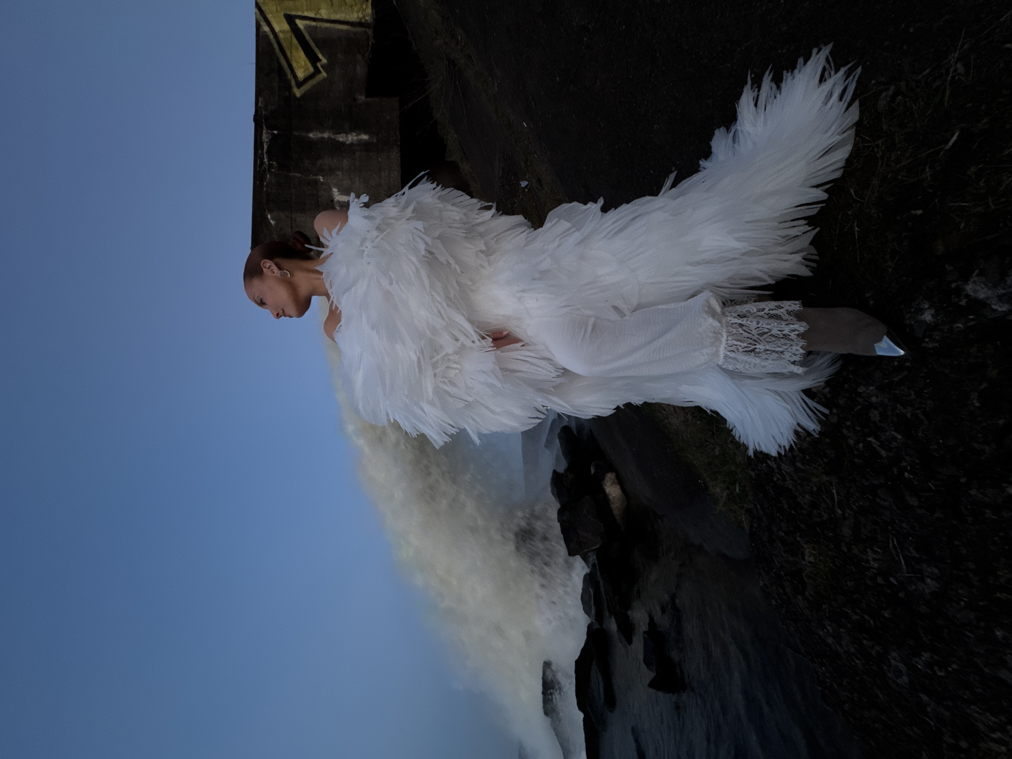 The photograph shows a woman in a white, feathered garment standing in an outdoor location, seemingly near a waterfall. The sky is visible in the background, indicating daylight. Part of a bridge or architectural structure appears in the upper left. The ground is dark and rocky. This is a photograph taken in or around the city of Chaikovsky, Russia.