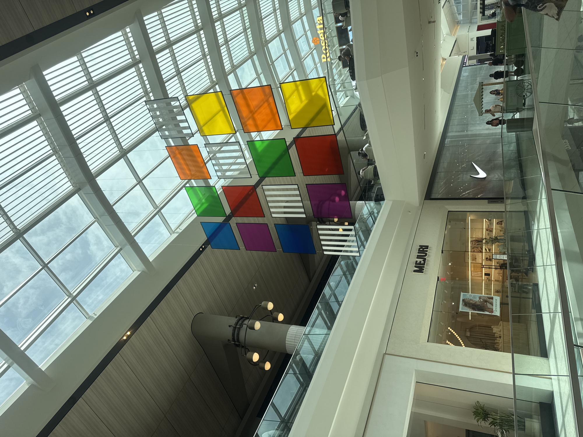 This media file captures an interior view of a modern shopping mall. The scene is dominated by a large, open atrium with a translucent, grid-like ceiling allowing natural light to filter in, suggesting daytime. Suspended from the ceiling is a colorful art installation composed of numerous square panes of translucent colored glass in hues of yellow, orange, green, red, blue, and purple, arranged in a geometric pattern.
Below the art installation, a cylindrical pillar with a circular light fixture is visible. The mall's architecture features clean lines, white and light-colored walls, and large glass panels. In the mid-ground, a store with a glass facade is identifiable by the prominent black lettering that reads "MEJURI". Within this store, displays of merchandise and products are visible, along with several people browsing. To the right of the "MEJURI" store, another storefront displays a large Nike logo.
Further back, other retail spaces and escalators can be seen, indicating a multi-level shopping environment. People are scattered throughout the mall, some standing, some walking, and some appear to be shopping or working. A small green vendor cart with an umbrella is visible near the "MEJURI" store. The overall impression is one of a bright, spacious, and contemporary commercial setting, likely a large shopping center or mall. There is no indication of specific weather conditions, and the emotions conveyed are neutral, focusing on the architectural and commercial aspects of the location.