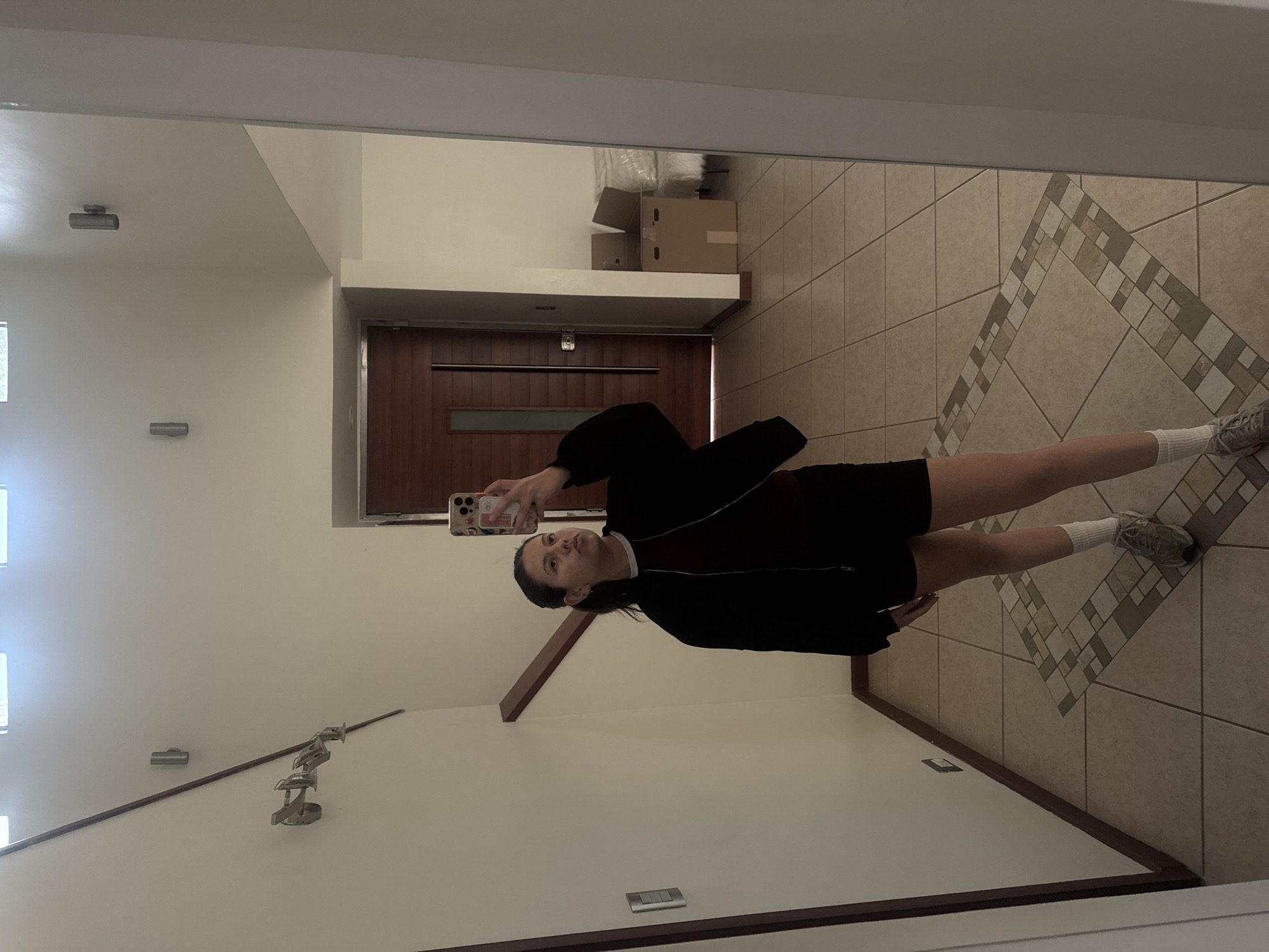 A mirror captures a woman in an indoor setting in San Pedro Cholula, Mexico. The woman holds a phone to take a picture of herself wearing a black jacket, shorts, and white socks with sneakers. The reflection shows a hallway with beige tiled floors featuring a decorative border pattern. A dark brown door is visible at the end of the hall, with boxes on a shelf above the door. The wall has light fixtures and a decorative coat rack. Daylight enters through a window.