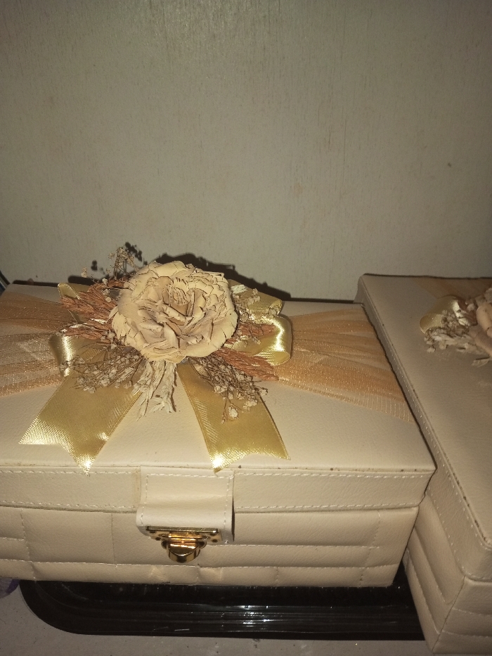 The image displays two elegant, rectangular boxes, likely designed for gifts or storage, set against a plain, light-colored wall. The most prominent box, positioned in the foreground and slightly to the left, is a light cream or beige color with a distinctive quilted or padded texture, featuring visible stitching lines. It is adorned with an elaborate decorative arrangement centered on its lid.
This decoration consists of a large, artificial or dried flower, possibly a rose, in a matching light beige hue, appearing to be made of delicate, layered material. Surrounding this central flower are clusters of dried, wispy elements: small, delicate white flowers (resembling baby's breath) and thin, brown, twig-like or straw-like pieces. Beneath and around this floral arrangement, a golden satin ribbon is tied into a bow, with its loops and tails extending outwards. Further accentuating the decoration is a wider, sheer, golden-colored ribbon or tulle, which is gathered around the base of the floral cluster, adding volume and shimmer. The front of this box features a small, gold-colored metal clasp, securing its lid.
To the right of the main box and slightly set back, a second, similarly styled box is partially visible. It shares the same light cream color and quilted texture as the first, suggesting it might be part of a set. A less elaborate, but matching, dried flower and ribbon decoration can be discerned on its lid.
Both boxes rest on a dark, reflective surface, possibly a tray or a polished tabletop. The background is a plain, light-colored wall, which exhibits some minor textural imperfections or dust marks, suggesting an indoor setting. The lighting is somewhat dim, creating soft shadows, particularly from the decorative elements onto the box lids and the wall behind them. The overall impression is one of delicate craftsmanship and elegant presentation, often associated with special occasion gifts or keepsakes. No text is visible in the image.