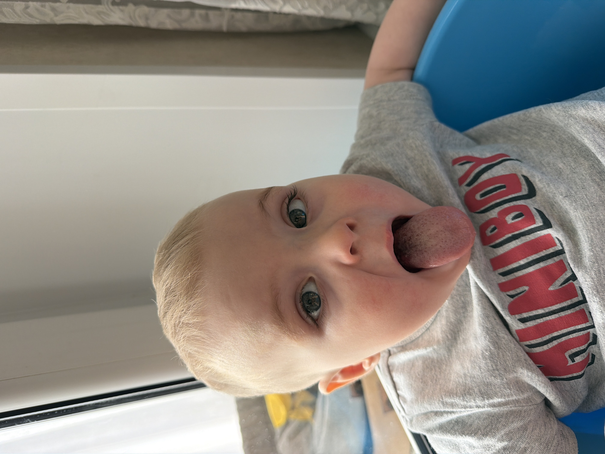 A toddler with light blond hair and blue eyes is shown in a close-up, upside-down view, sticking their tongue out. The toddler is wearing a grey shirt with a red and black design. The toddler is sitting in a blue chair. The scene appears to be indoors, next to a window. It is impossible to determine the location in the city of Votkinsk, Russia from the image.