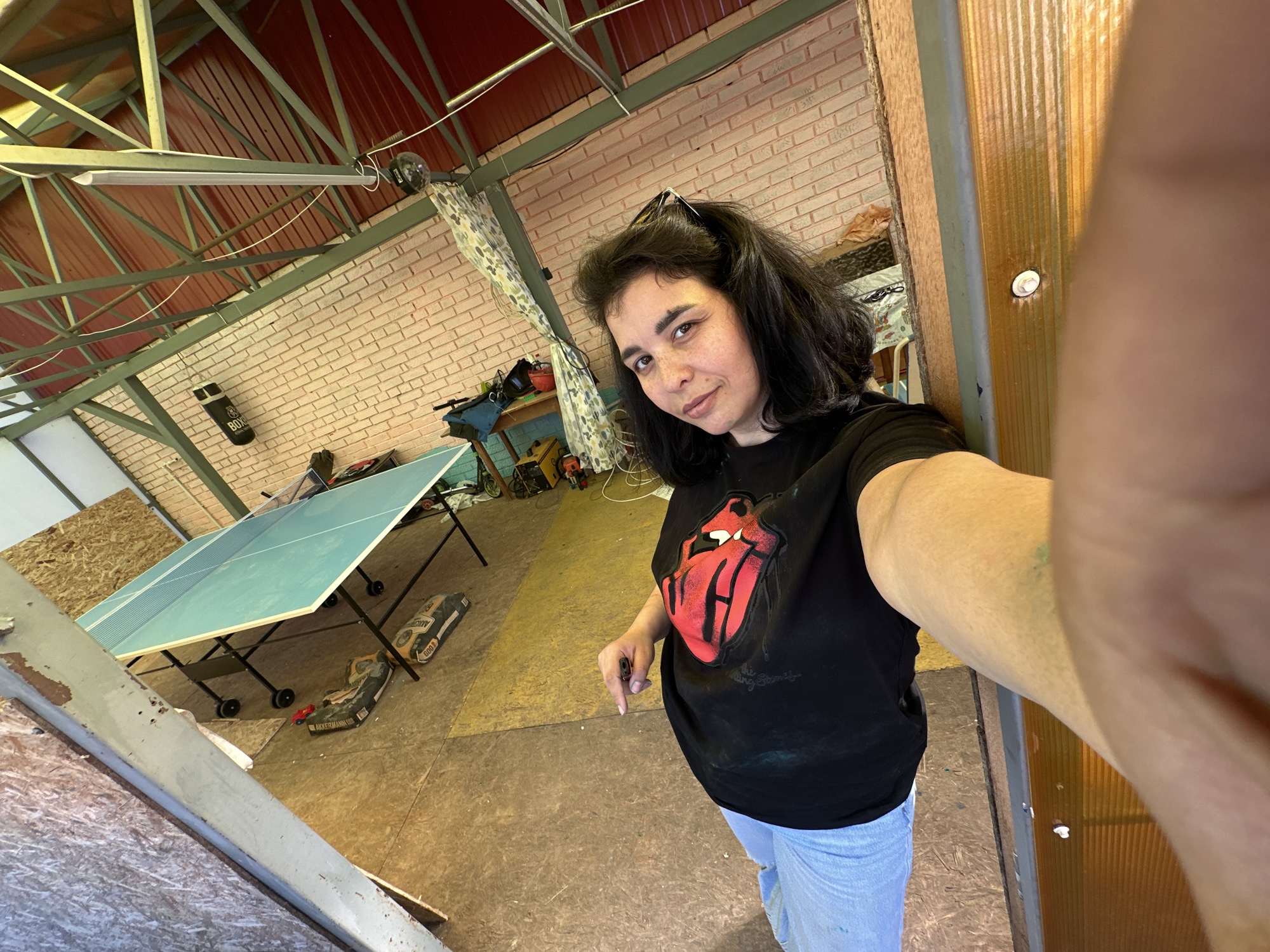 A woman takes a selfie in a building with a red metal roof supported by a steel frame. The woman has dark hair, light skin, and wears a black t-shirt with a red lips graphic and light blue jeans. She holds a pen in her left hand. A ping pong table stands in the background. The floor is plywood. The walls consist of unfinished brick and wood. A curtain hangs by the brick wall. There are tools and other items scattered around, including boxes on the floor.