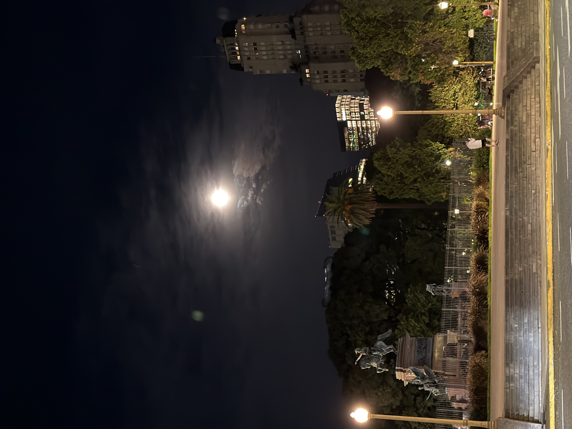 The image shows a nighttime scene in Buenos Aires, Argentina. The full moon is visible in the dark sky, partially obscured by clouds. A row of buildings lines the right side of the frame, interspersed with trees and illuminated by street lamps. Some people are visible on the right side of the image, walking along a path near the edge of the street. The road has a yellow line. There are sculptures on the left side.