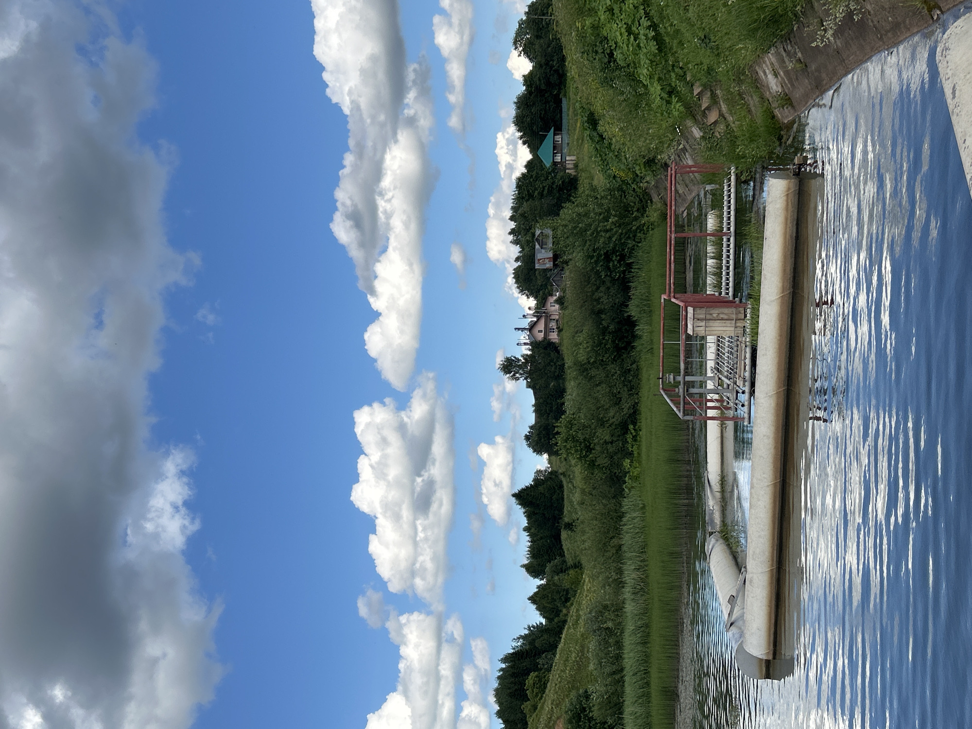 In the city of Verkh-Rozhdestvo, Russia, a body of water is visible, likely part of the settlement's water management system. In the foreground, large, white pipes float on the surface, partially submerged. Adjacent to these pipes is a metal grate structure with red railings, situated on a platform within the water. The water itself is calm, reflecting the blue sky and white clouds that dominate the upper portion of the image. Beyond the water, a bank of tall green grass and reeds lines the edge, leading to a gently sloping hill covered in lush green trees. In the distance, several houses are nestled amongst the trees, some with visible roofs of various colors, including pink and green. The overall scene depicts a rural landscape with elements of industrial infrastructure integrated into the natural environment.