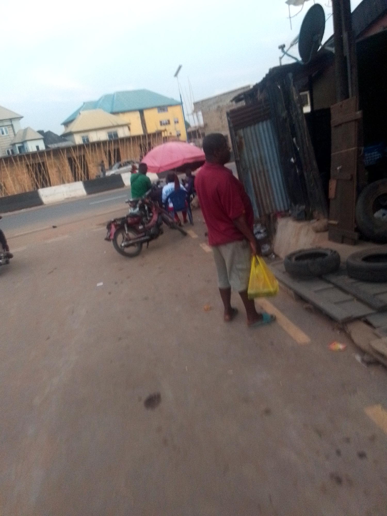 The image depicts a street scene. A man in a red shirt and khaki shorts stands on the right side of the image, holding a yellow plastic bag. To his right, there's a makeshift structure or stall with a satellite dish on top, and some tires are visible near the stall. A motorcycle is parked on the left side of the image with other people gathered under a pink umbrella. In the background, there are buildings and a barrier along the road. The location appears to be in the city of Unknown, Unknown.