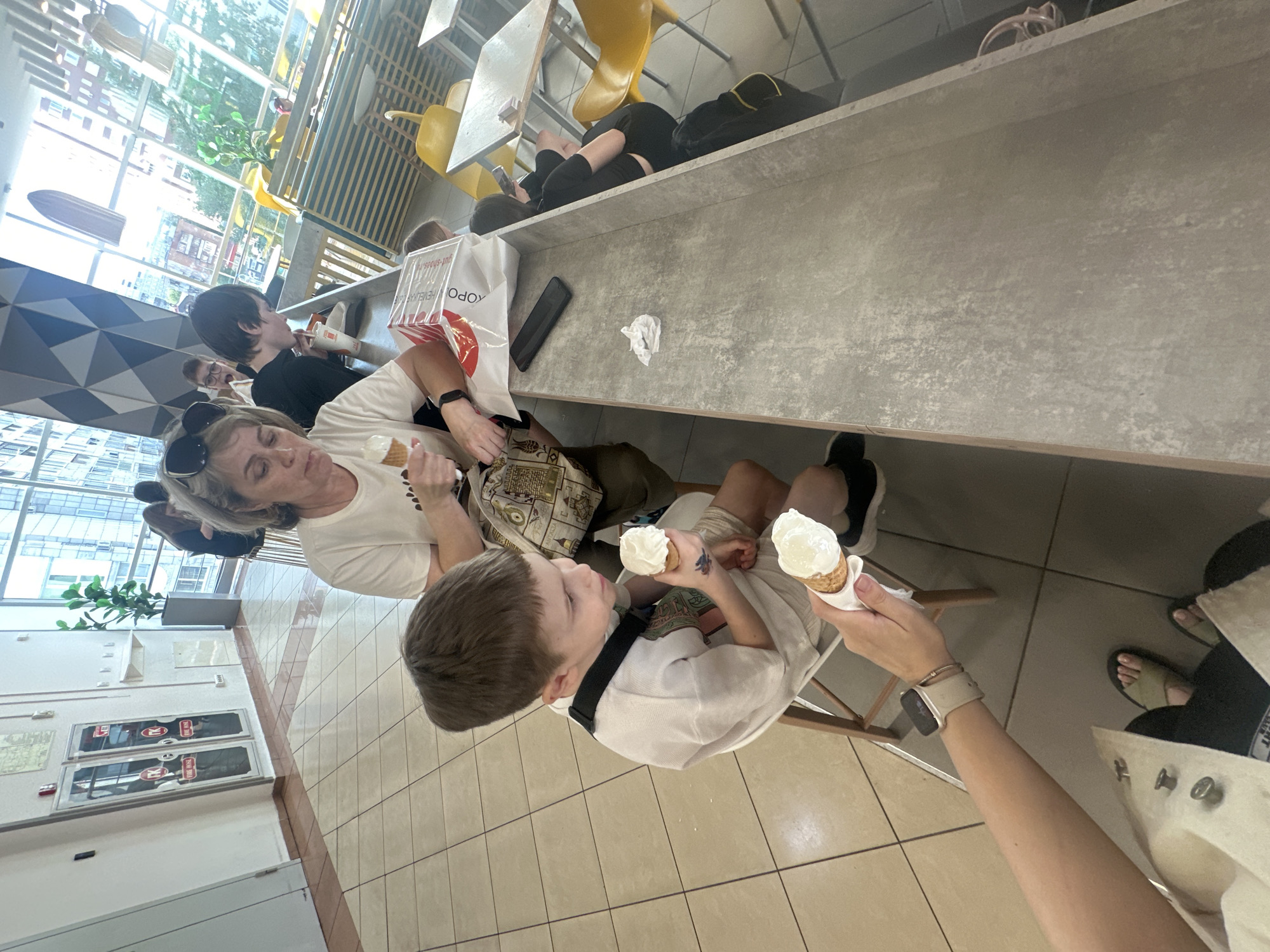The image shows a woman and a boy eating ice cream in a brightly lit indoor space, likely a cafe or restaurant. Another person is visible in the background, as well as someone lying on the countertop. The space appears to be in Perm, Russia, judging from a shopping bag in the image with Cyrillic text.