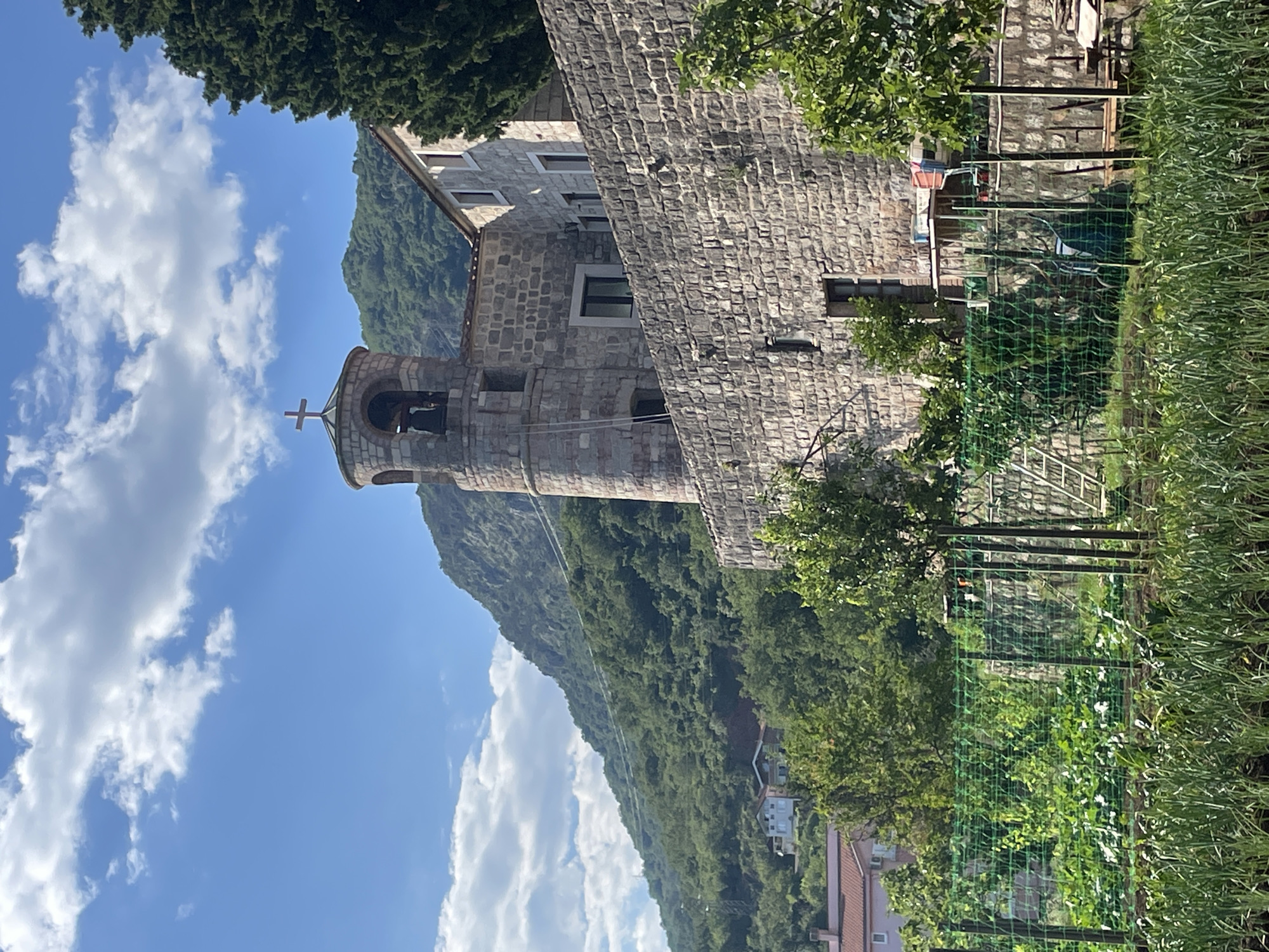 The image shows a stone building, likely a church or monastery, with a bell tower topped by a cross, against a backdrop of a mountain and a blue sky with clouds. The building is constructed of stone blocks. To the right, a garden area is visible with a fence. The location is likely in the city of Budva, Montenegro, given the style of architecture and the landscape.