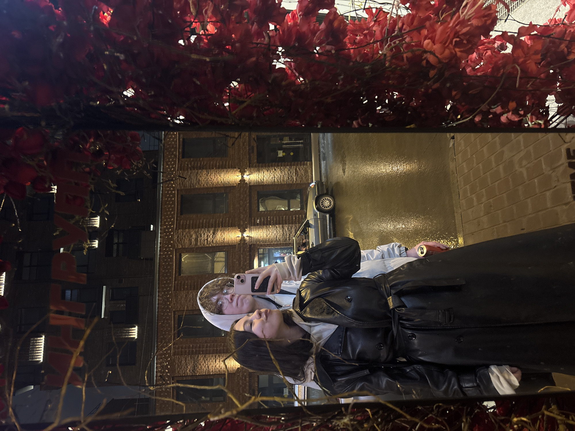 Two people are reflected in a surface with red foliage above. One person is holding a phone and taking a photograph of the other. The reflection shows a building with lit windows, a vehicle, and a wet street. The location is Perm, Russia.