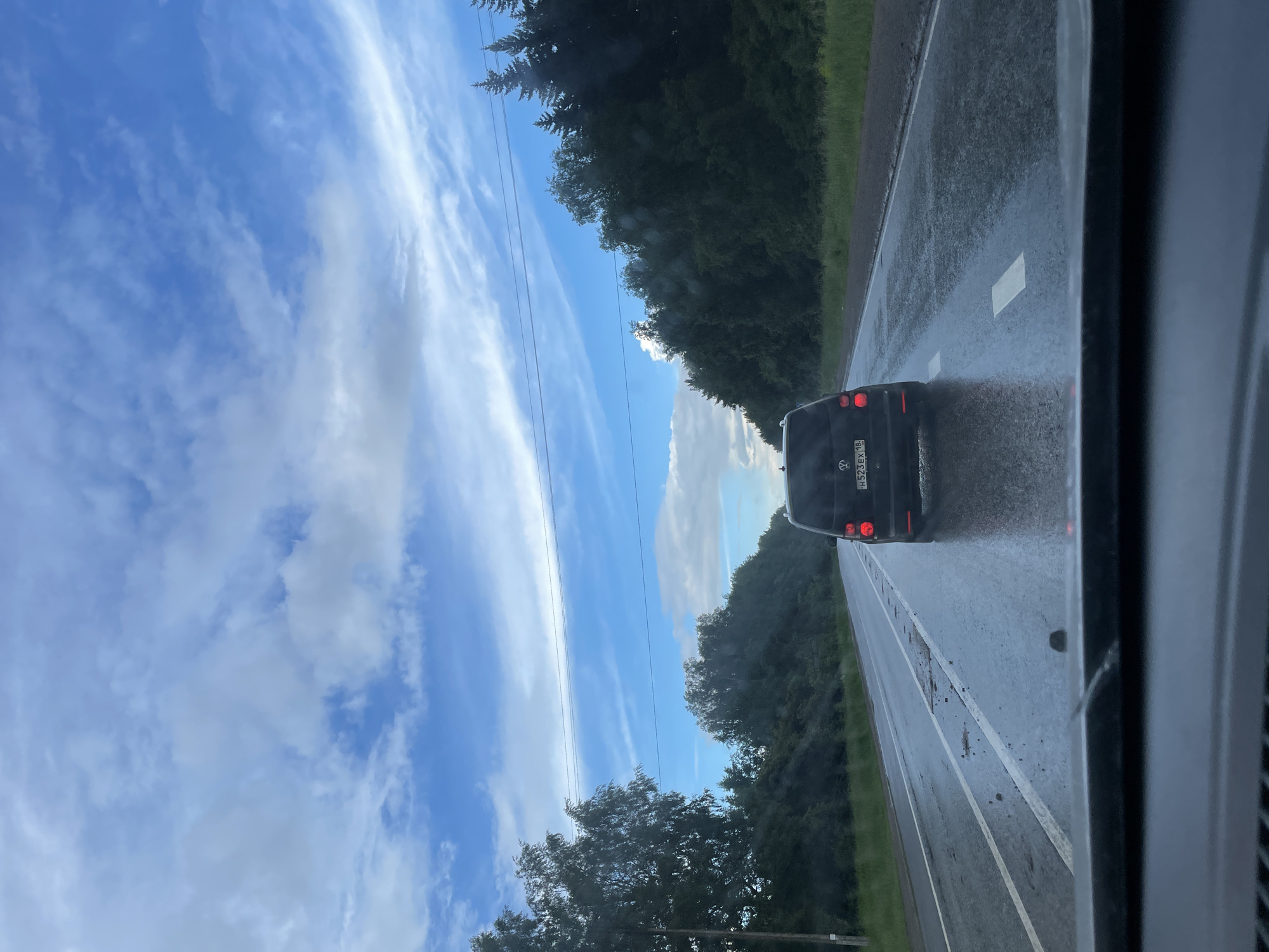 A black Volkswagen Touran with a license plate reading H523EX 16 is driving on a wet, asphalt road. The road has white lane markings and some debris. The sky is blue with scattered white clouds. Trees line the side of the road. The image appears to be taken from another vehicle driving behind the Volkswagen. The location is identified as Dvigatel', Russia.