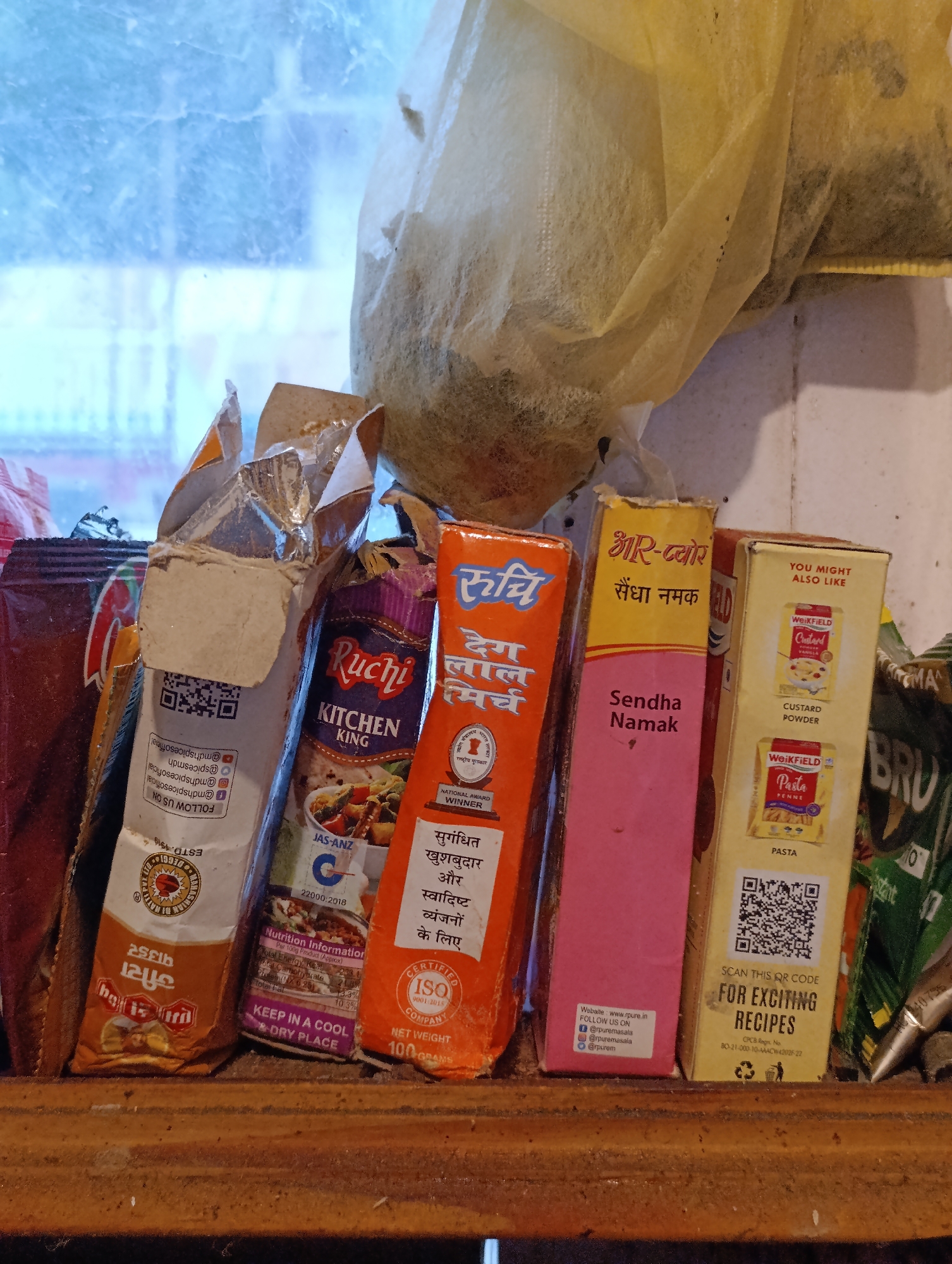The image shows a shelf in what appears to be a shop or kitchen in Vashisht, India, displaying various food products. The products are primarily packaged spices and cooking ingredients. From left to right, there is a dark brown packet with red and white lettering, partially obscured. Next to it is a white cardboard box with silver foil lining and some text, including a QR code. Following this is a purple and blue packet labeled "Ruchi Kitchen King." To its right is an orange-colored box with Hindi text and a "Ruchi" logo, featuring "Degh Lal Mirch" and a national award winner emblem. A pink box with Hindi text and "Sendha Namak" is to the right of the orange box. Finally, a yellow box labeled "WEIKFIELD Custard Powder" and "WEIKFIELD Pasta Penne" is on the far right, also containing a QR code and offering "FOR EXCITING RECIPES." The shelf itself is made of rough, dark wood. The background shows a window with a light blue, possibly frosted or textured glass, and a pale yellow draped material.