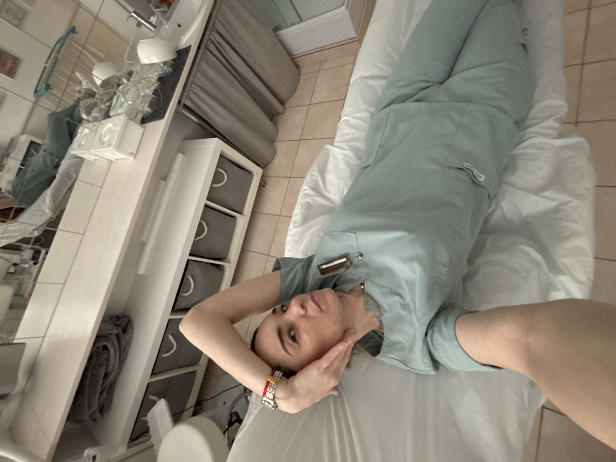 A medium shot captures a woman in light blue scrubs lying on a white treatment table. She is angled slightly, with her head positioned near the left side of the frame and her body extending towards the upper right corner. Her hands are near her face. She wears a bracelet on her left wrist. Her name badge is visible on her left chest.
The background features a clinical setting with a white cabinet and storage drawers along the left side. A sink area with various containers is partially visible. The floor has light beige tiles. This photo was not taken in Romanovka, Russia.