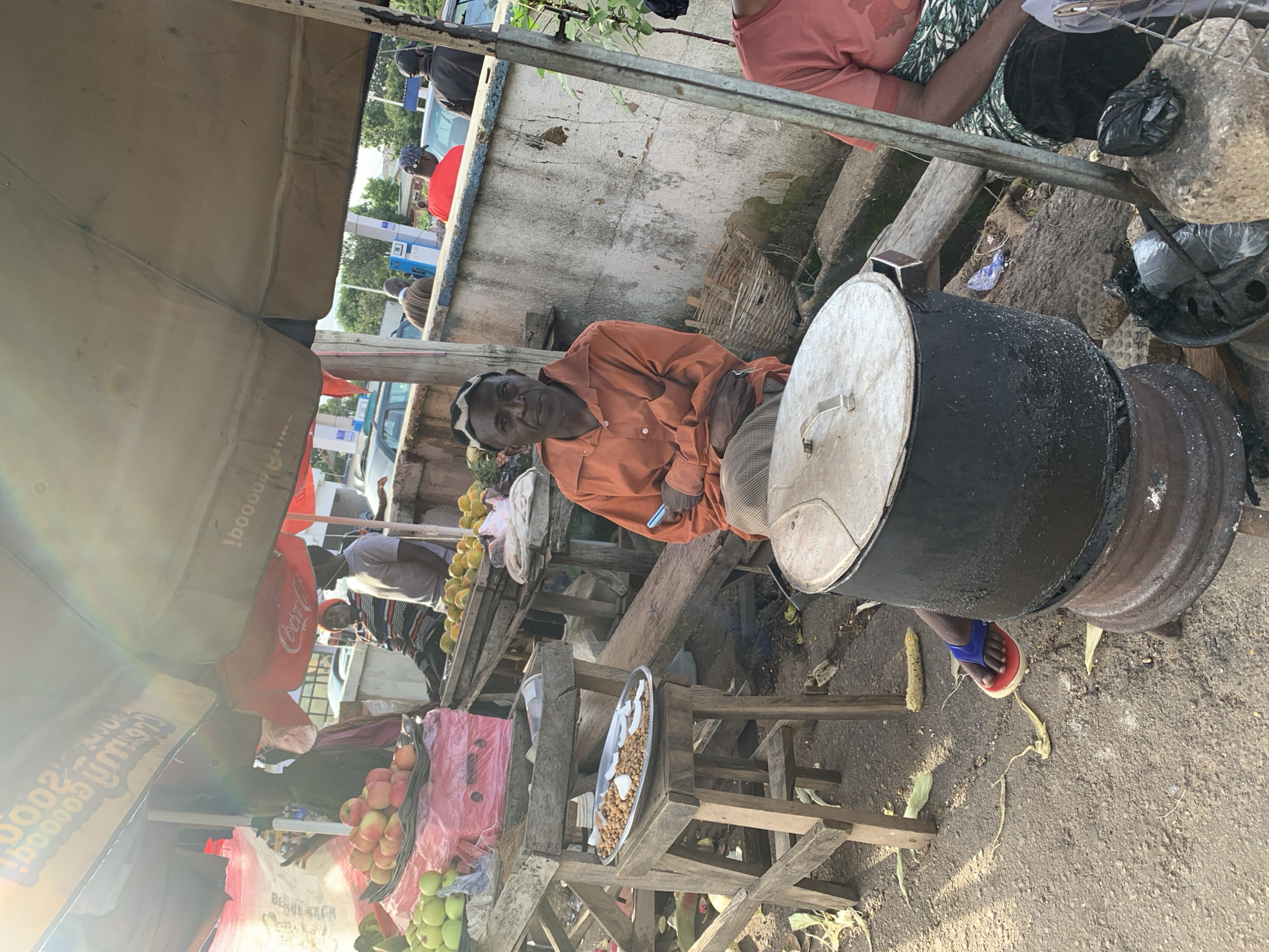 This image depicts an outdoor market scene in Jos, Nigeria. A man in an orange shirt sits beside a large cooking pot, holding a blue object, possibly a phone. In front of him, on a wooden stool, is a metal tray with food items. To his left, a stall displays apples and green fruits. Further back, other vendors and customers are visible, with cars and a fuel station in the background. The environment appears to be a busy roadside market, with a mix of food stalls and everyday commerce.