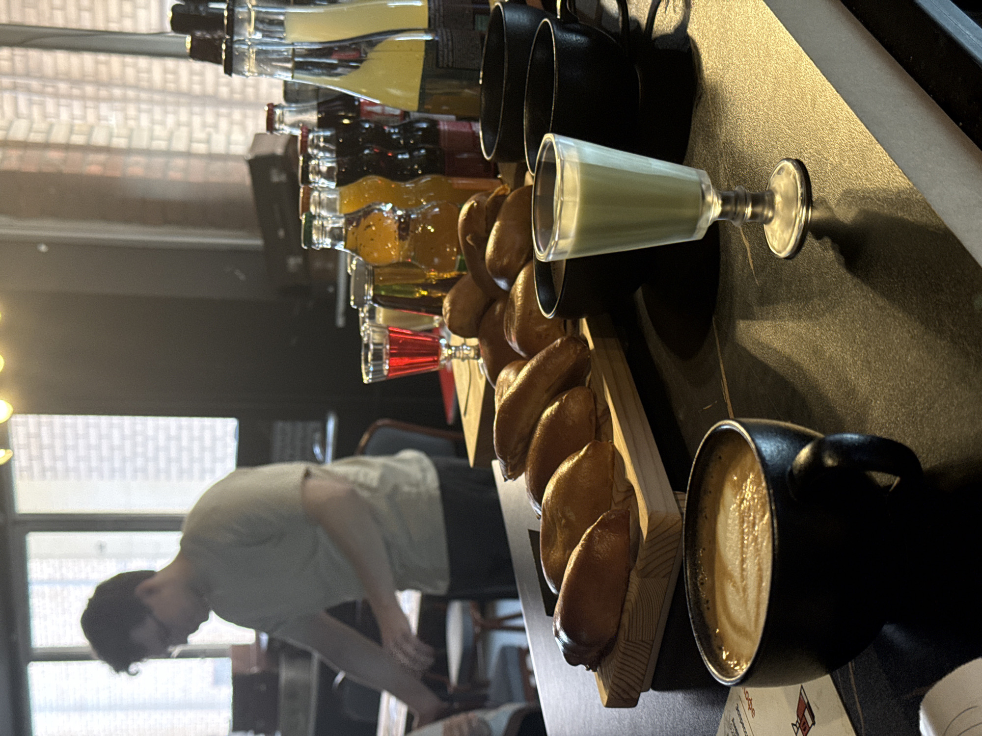 The image shows a counter or bar area in what appears to be a café, possibly in Moscow, Russia, based on the Cyrillic text visible on a menu partially visible at the bottom right of the frame. A man in a light-colored t-shirt is leaning towards the left, seemingly attending to something out of frame. On the counter, there's a row of bottles filled with various colored liquids, potentially syrups or juices, lined up behind black mugs and a stemmed glass containing a green substance. A wooden rack displays a row of pastries, likely buns or rolls. A mug filled with coffee sits on the right side of the frame, and the background includes a window with a view of what looks like another building or cityscape. The lighting is somewhat dim, but focused on the counter area.