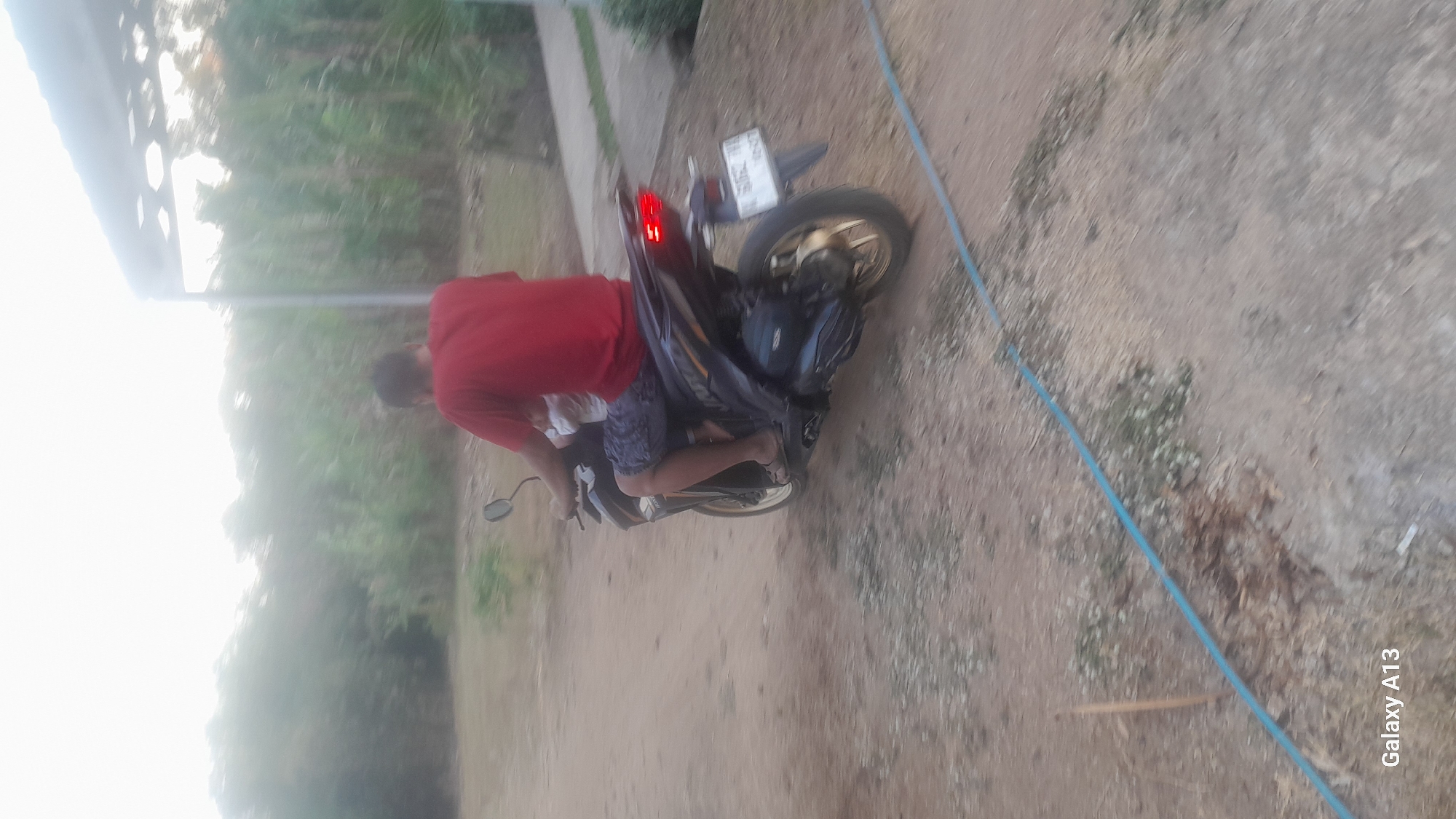 A person wearing a red t-shirt and patterned shorts is riding a dark-colored motorcycle. Another person, possibly a child, is seated in front of the rider. The motorcycle has a license plate that is partially obscured by motion blur. The rider's left foot is resting on the ground. The motorcycle is on a dirt road with sparse vegetation and some scattered debris. In the background, there are trees and a structure with a roof. A blue hose is visible on the ground in the foreground. The image also contains a watermark that says "Galaxy A13". This scene appears to be in a rural or semi-rural area.