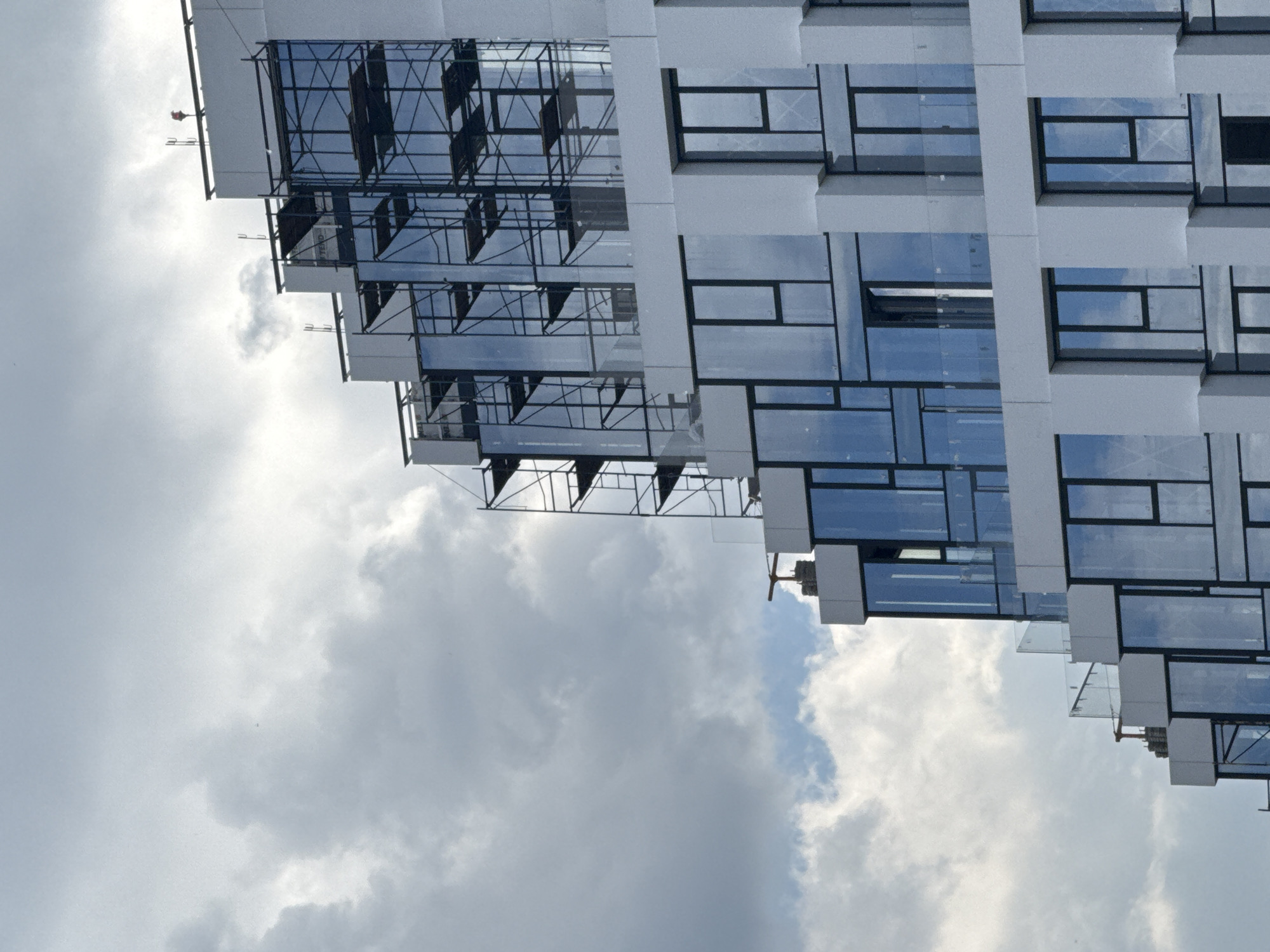 This image captures a modern, multi-story building with a prominent glass facade, set against a cloudy daytime sky. The building features numerous rectangular glass windows framed by light-colored, possibly white or light gray, exterior panels, creating a grid-like pattern. Its left side exhibits a stepped or terraced design, where extensive scaffolding is erected, suggesting ongoing construction or maintenance work.
The sky is largely overcast with significant white and gray clouds, though patches of lighter sky, possibly indicating thinner clouds or hints of blue, are visible, particularly reflected in the building's windows. These reflections also show the intricate structure of the scaffolding.
On the very top edge of the building, near the scaffolding on the left, a small, distinct red object is visible, possibly a person (a construction worker) or a piece of equipment. No clear activity is discernible beyond the general presence of the scaffolding.
Very faint, distorted text, possibly reading "TTEND" or similar, can be seen reflected in one of the glass panels on the left side of the building, though it is not clearly legible. The image is oriented sideways, with the building extending diagonally from the lower right to the upper left. This scene is located in Moscow, Russia.