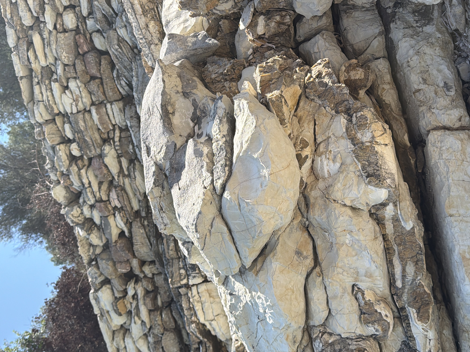 The image shows a close-up view of layered rocks, possibly forming a natural cliff face or an old stone wall, under a clear blue sky. The rocks vary in color from light beige and white to tan and brown, with some darker veins and textures. The foreground is dominated by large, irregularly shaped rocks with visible cracks and fissures. In the background, more layers of smaller, roughly stacked stones are visible, with some sparse vegetation and trees at the very top. The lighting suggests it is daytime, likely midday, with sunlight casting shadows and highlighting the rugged surfaces of the rocks. The scene is devoid of any people or animals. This geological formation is located in the Municipal Unit of Kastos, Greece.